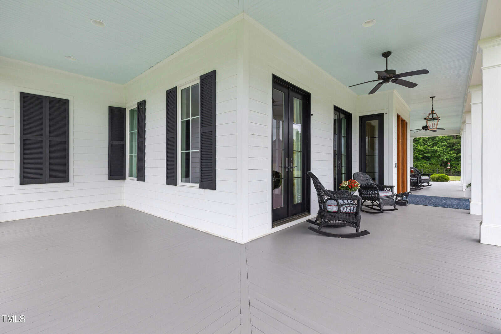 A covered porch with a light blue ceiling features black rocking chairs, a ceiling fan, black shutters on white siding, and a view of greenery in the background.