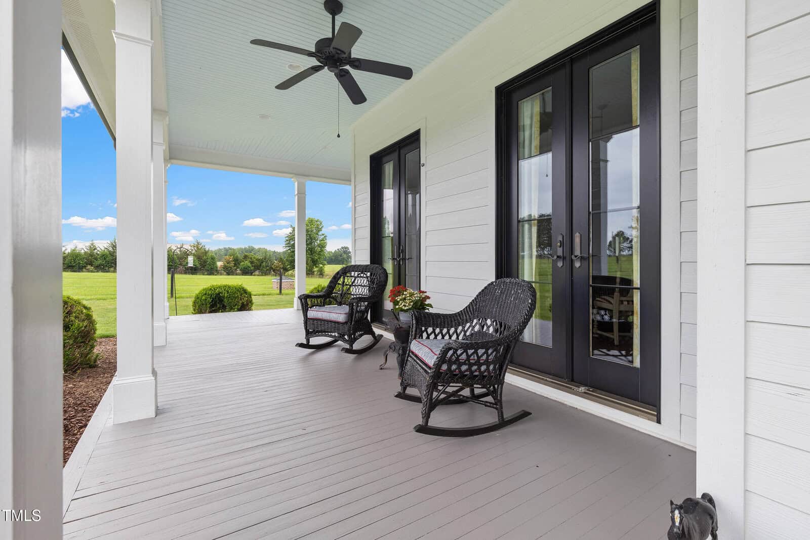 A covered porch with two black wicker rocking chairs, a small table with a plant, ceiling fan, double glass doors, and white siding. The porch overlooks a green lawn and open landscape under a blue sky.