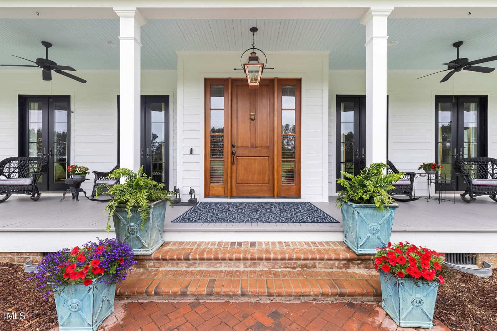 A house entrance with a wooden double door, brick steps, blue potted plants with flowers, a blue doormat, white porch columns, ceiling fans, and black outdoor furniture on a covered porch.