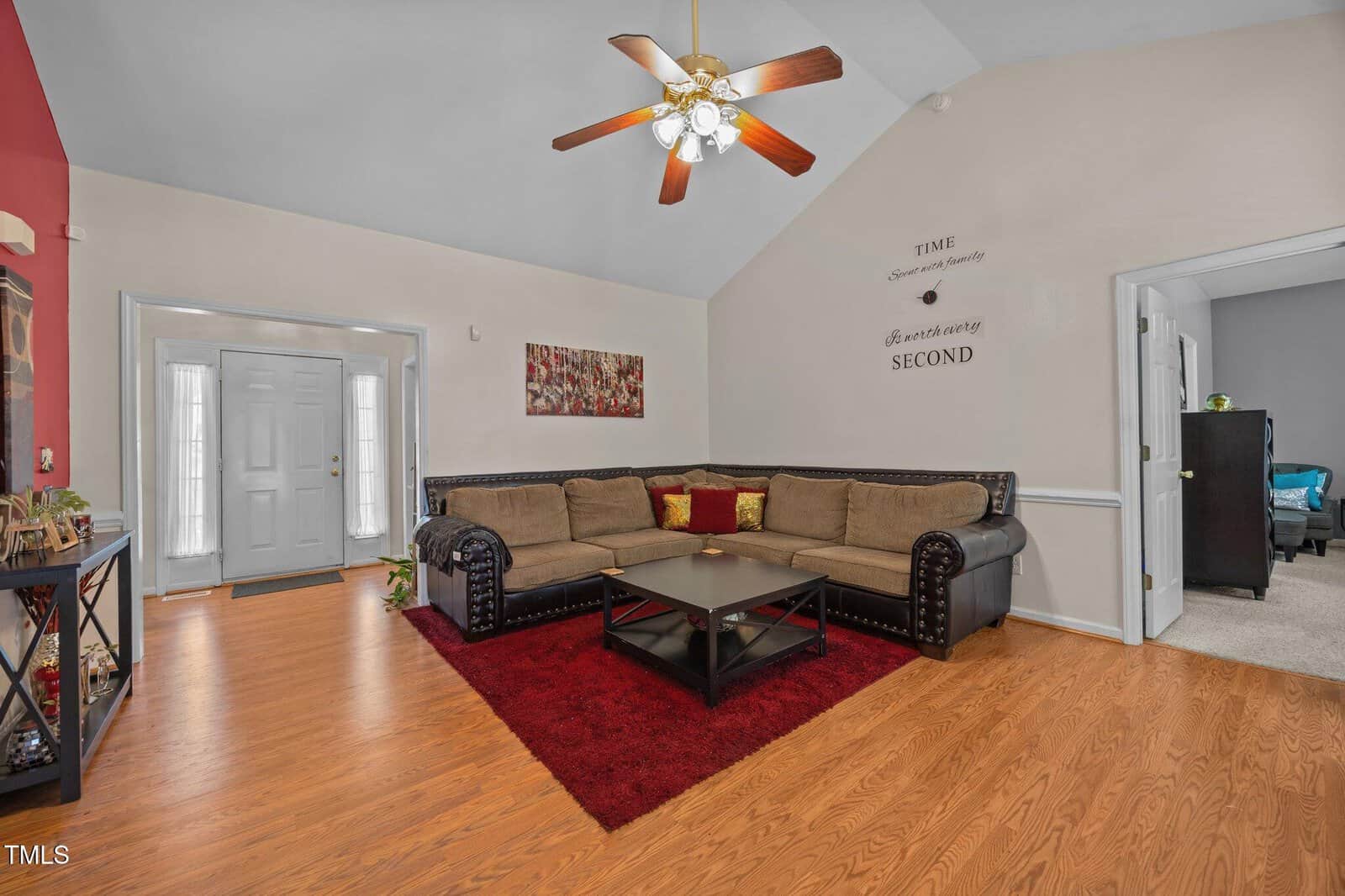 A living room with wood flooring, a brown sectional sofa, a black coffee table on a red rug, wall art, and a ceiling fan with lights. An entryway and a doorway to another room are visible.