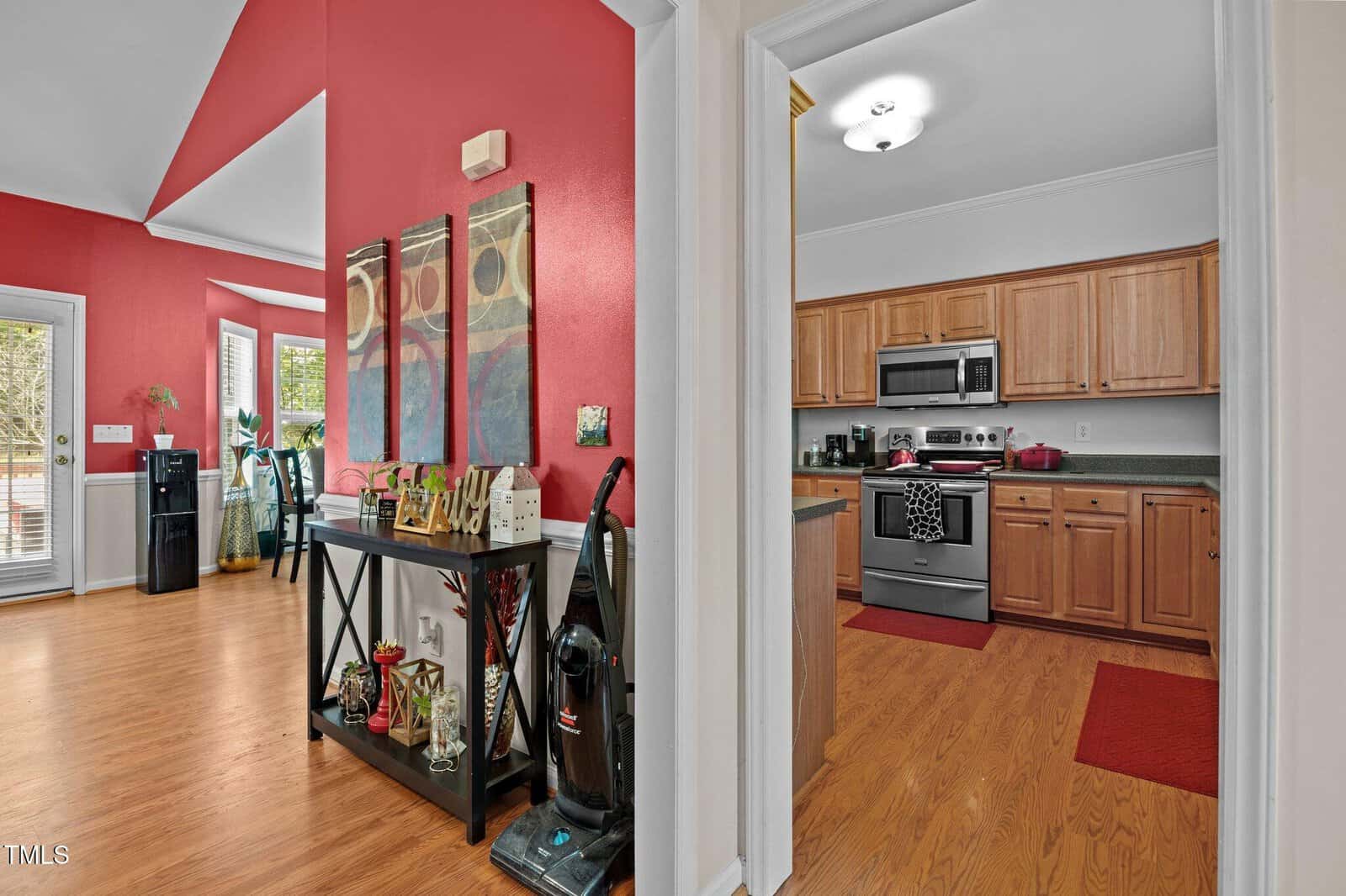 A kitchen with stainless steel appliances and wooden cabinets is visible through a doorway. The adjacent room has red walls, a drink station, a vacuum cleaner, and large windows letting in natural light.