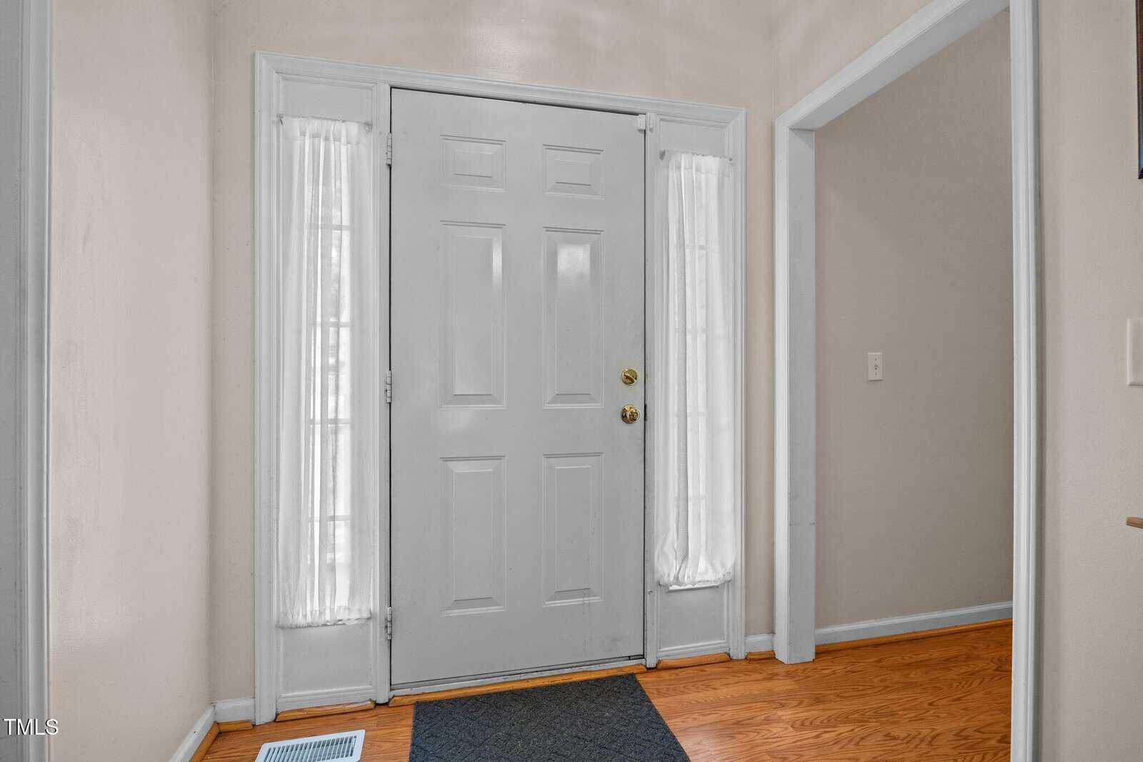 A white front door with two narrow windows on each side covered by sheer white curtains, set in a beige entryway with hardwood floors and a black doormat. An open doorway leads to another room.