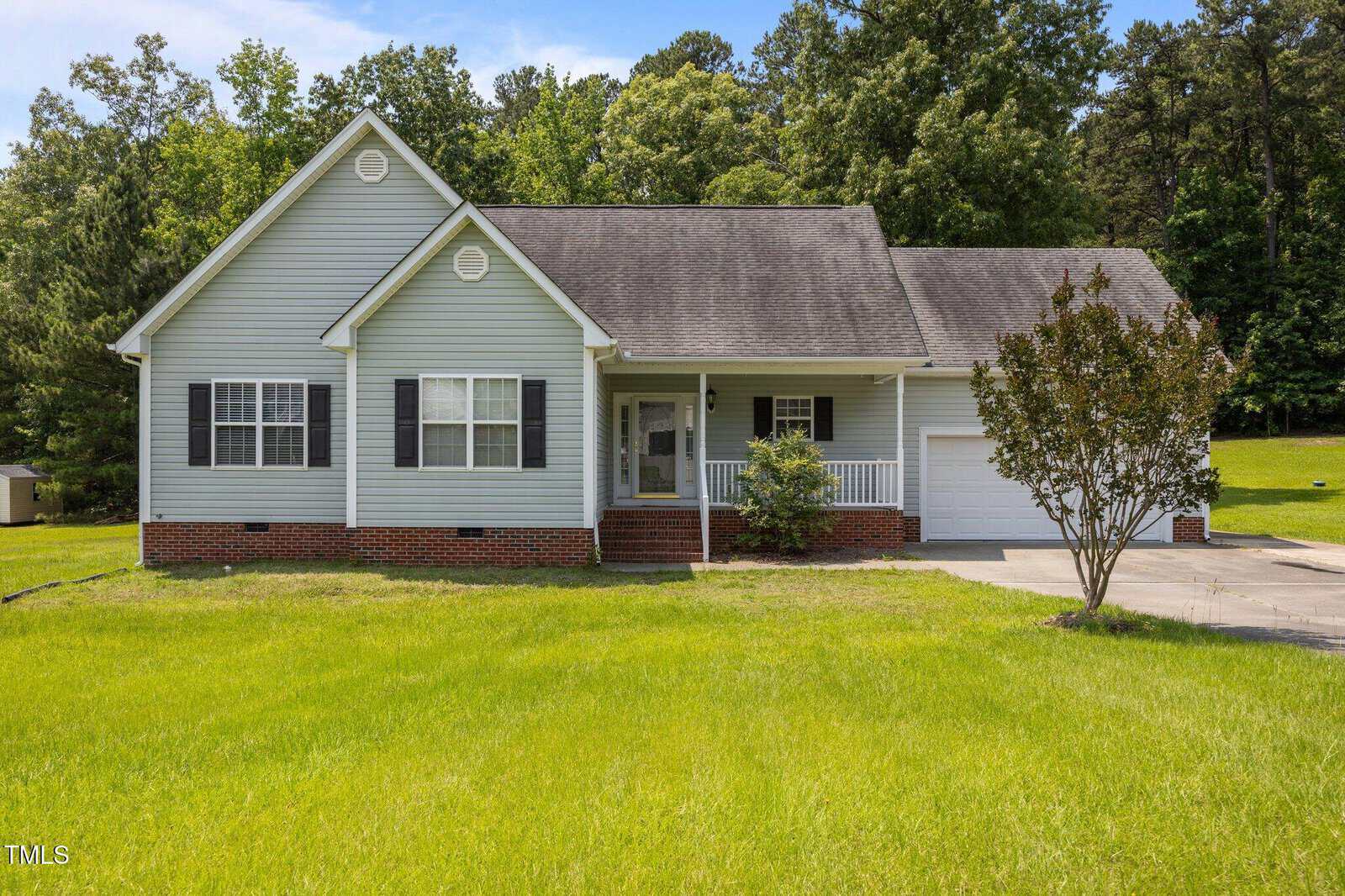 Single-story house with light gray siding, black shutters, and a covered front porch. There is a one-car garage, a concrete driveway, and a lawn with a small tree and bushes. Tall trees are in the background.