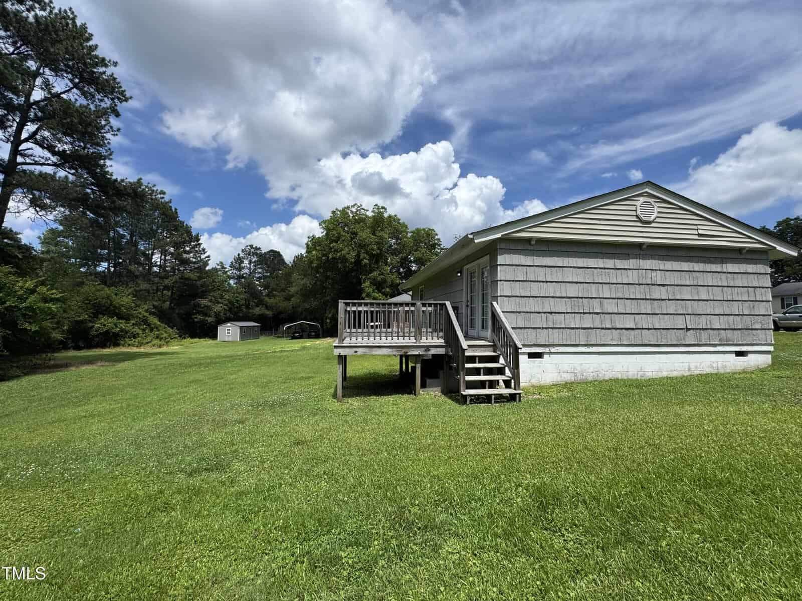 A single-story house with gray siding and a small wooden deck sits on a large grassy lawn. Trees and a small shed are visible in the background under a partly cloudy sky.