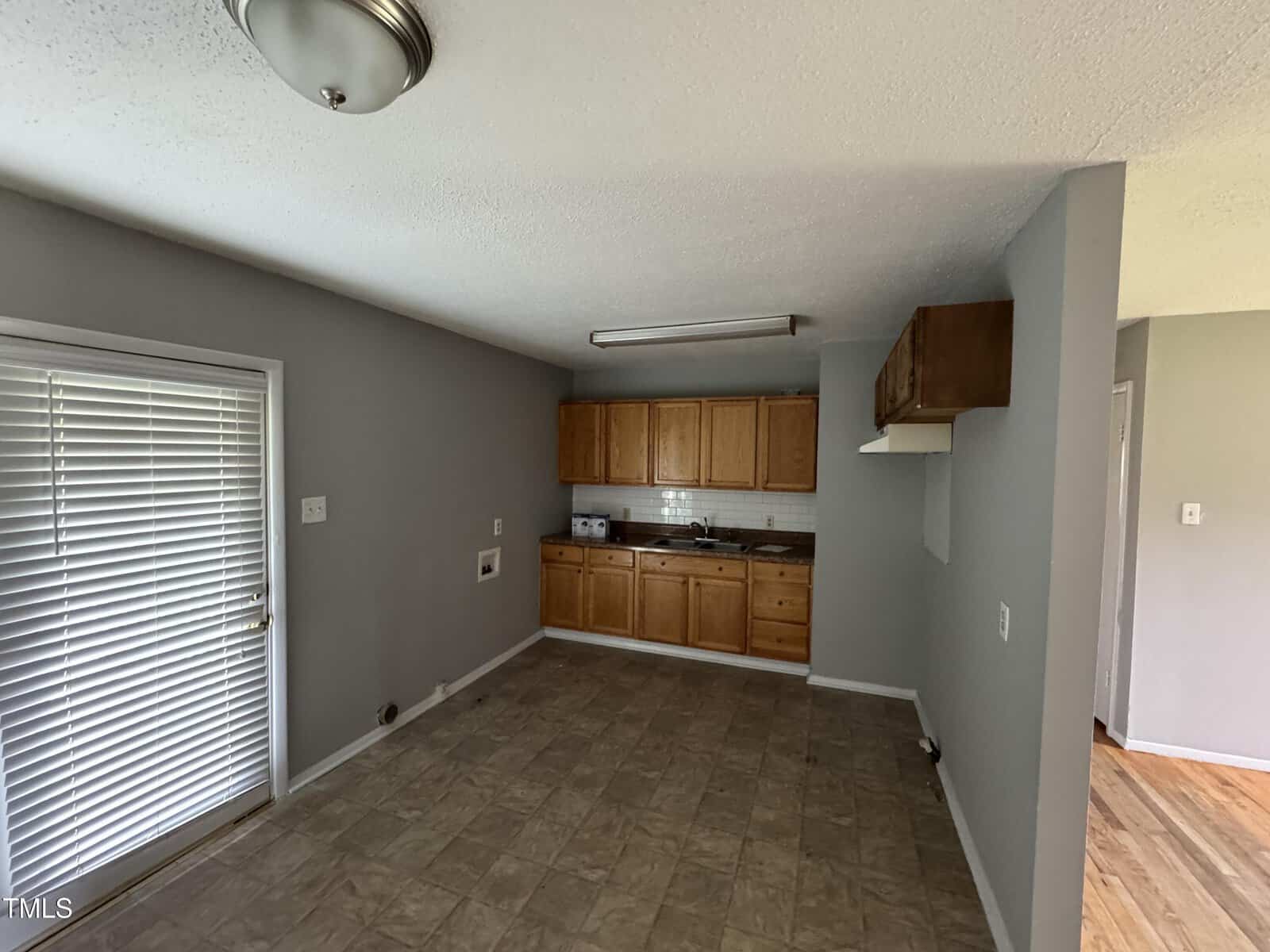 Small kitchen with wooden cabinets, light gray walls, and a tiled floor. There’s a window with blinds and a door on the left, and a bright overhead light fixture on the ceiling.