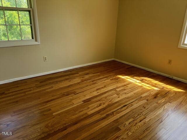 Empty room with wood flooring, cream-colored walls, and two windows letting in natural light. Sunlight forms a bright patch on the floor near one window. No furniture or decorations present.