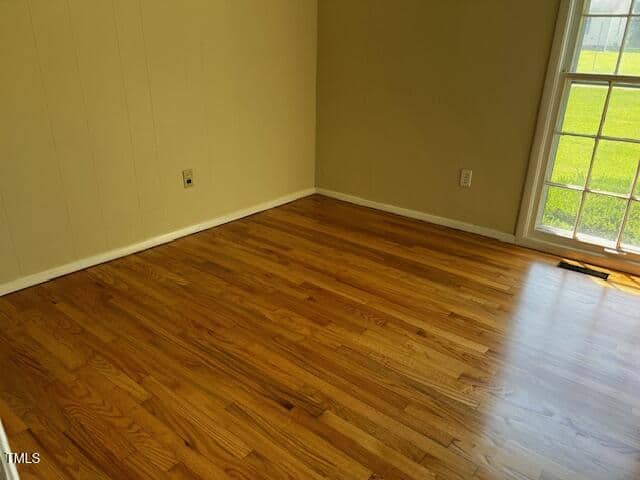 Empty room with light-colored painted walls, hardwood flooring, and a large window letting in natural light. The window overlooks a green lawn outside.