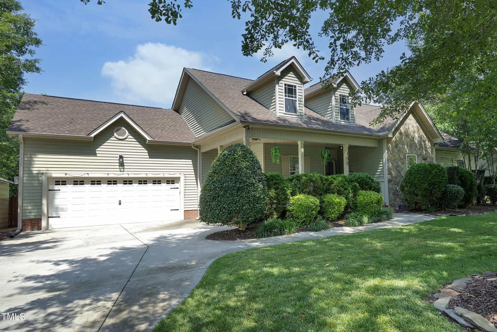 A beige suburban house with a double garage, dormer windows, and a well-manicured lawn and shrubs, viewed on a sunny day with clear blue skies and some shade from nearby trees.