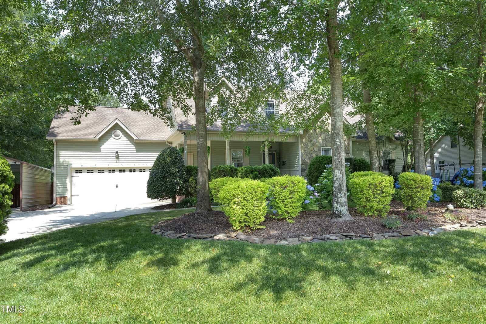 Single-story house with a two-car garage, covered front porch, and beige siding. The front yard features manicured grass, neatly trimmed bushes, landscaping stones, and several tall trees providing shade.