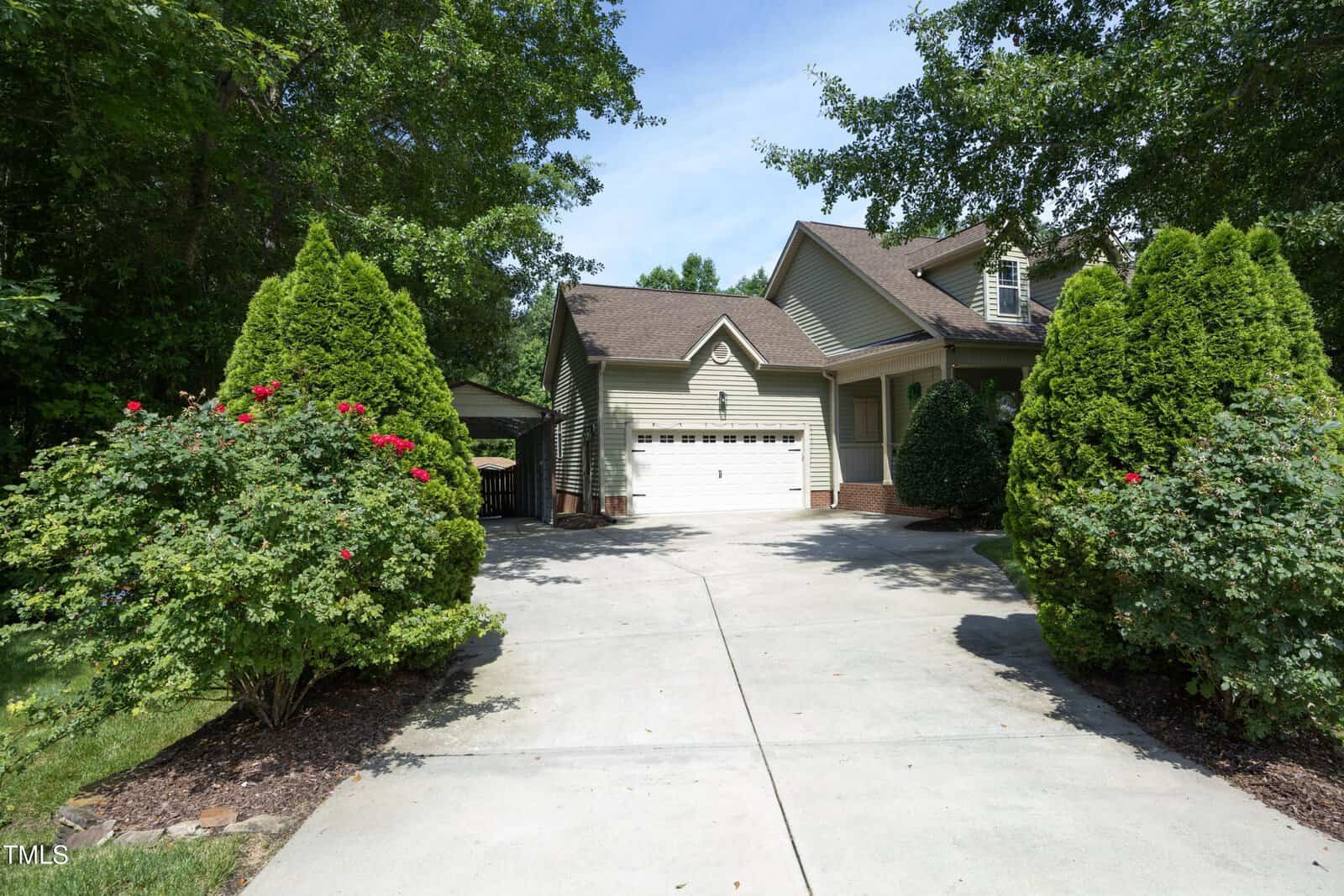 A suburban house with a two-car garage, light-colored siding, and a gabled roof. The driveway is bordered by neatly trimmed bushes and small trees, with surrounding greenery and clear blue sky.