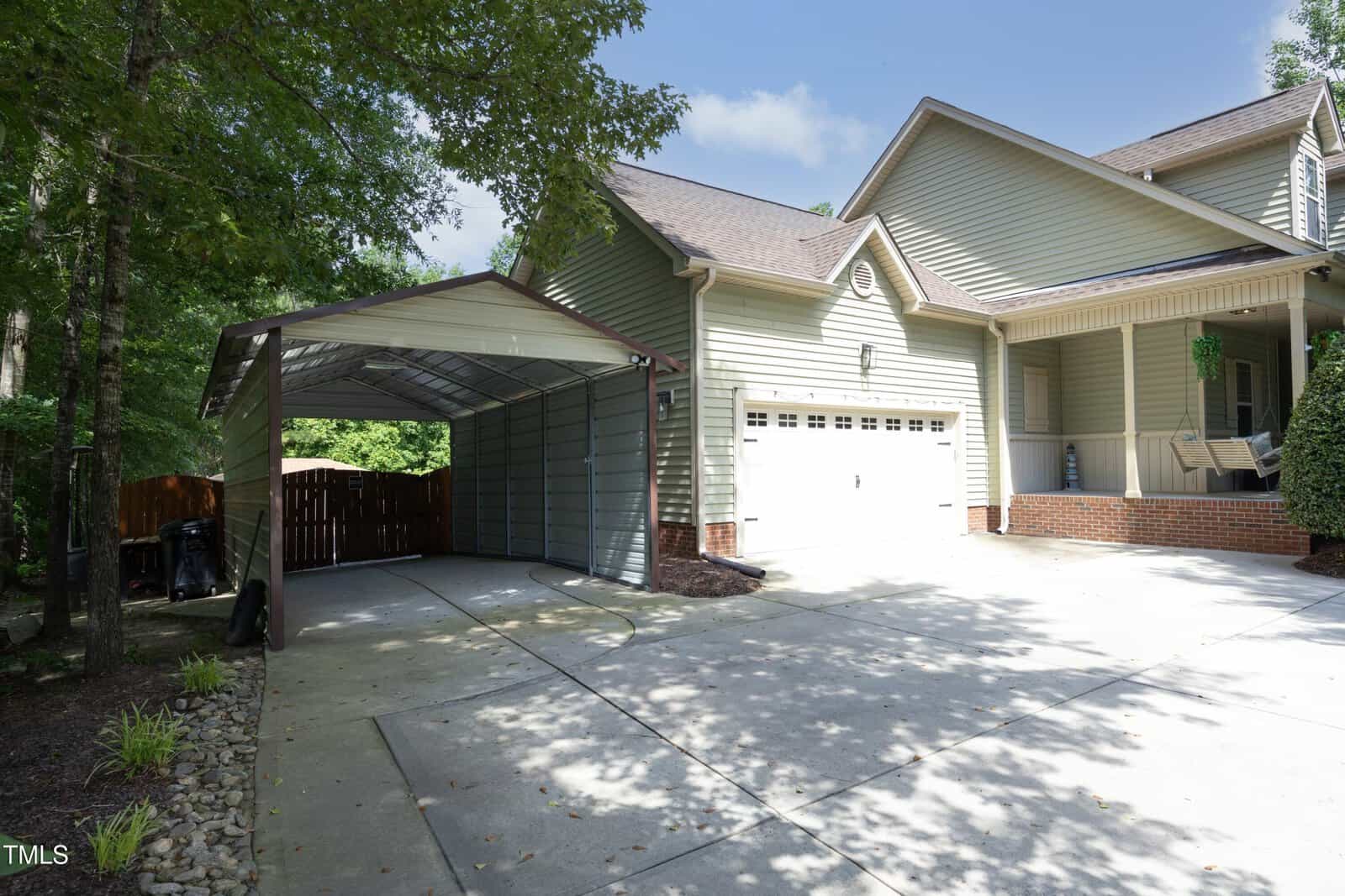 A light green house with a two-car garage and a covered carport sits on a spacious concrete driveway, surrounded by trees and landscaping with rocks and mulch.