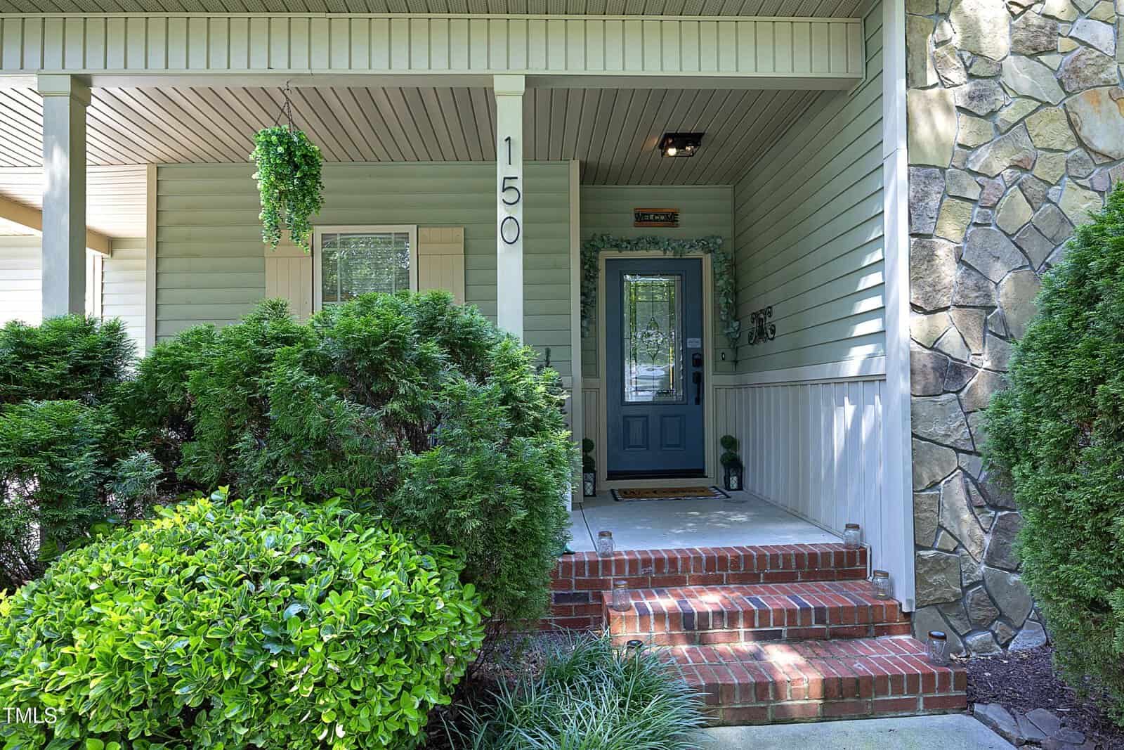 A house entrance with brick steps, a blue front door, the number 150 displayed, leafy bushes, hanging plant, and a stone and siding exterior.