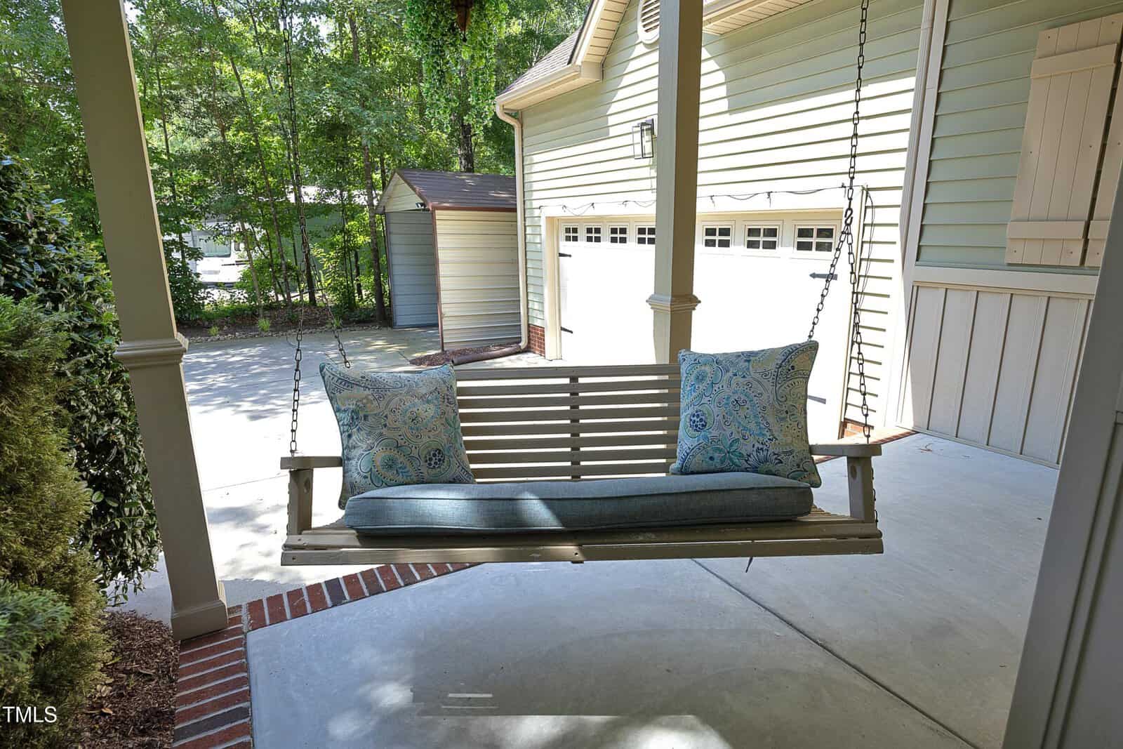 A wooden porch swing with blue cushions and two patterned pillows hangs on a covered patio. A driveway, a detached garage, and trees are visible in the background.