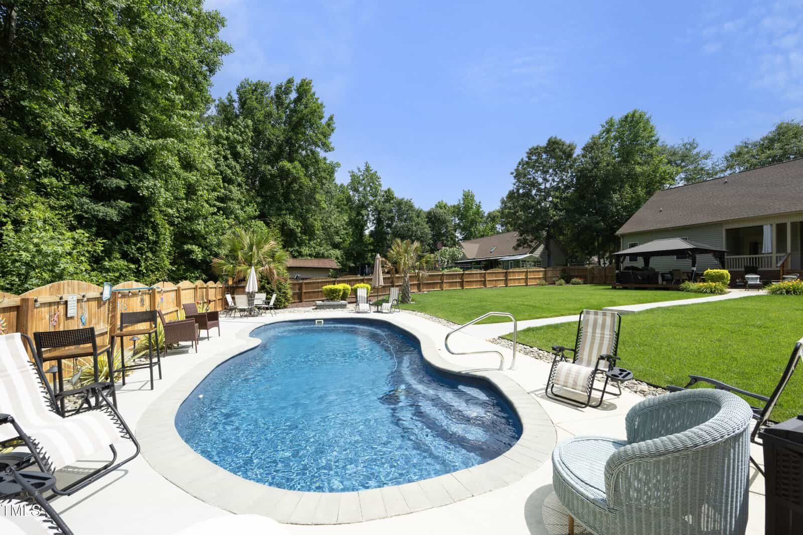 Backyard with an in-ground swimming pool surrounded by a concrete deck, lounge chairs, and patio furniture. The yard has green grass, a wooden fence, and trees along the perimeter. Houses visible in the background.