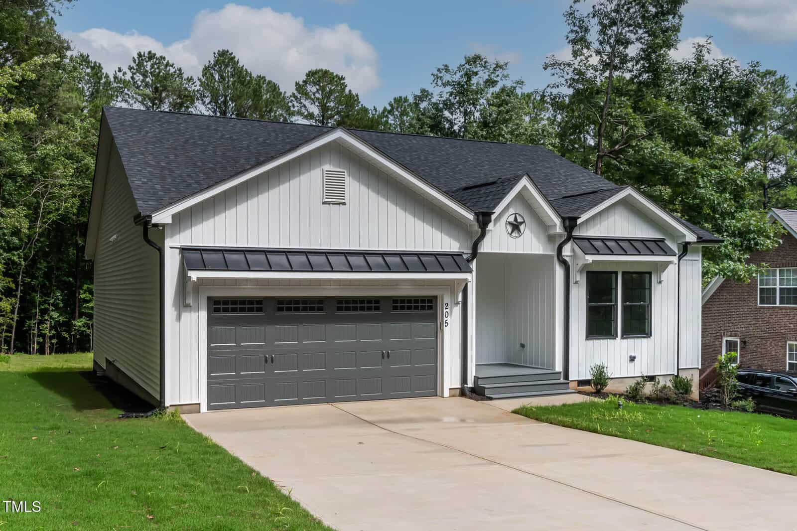 A modern single-story white house with a gray double garage door, black roof, and a small front porch, surrounded by grass and trees under a partly cloudy sky.