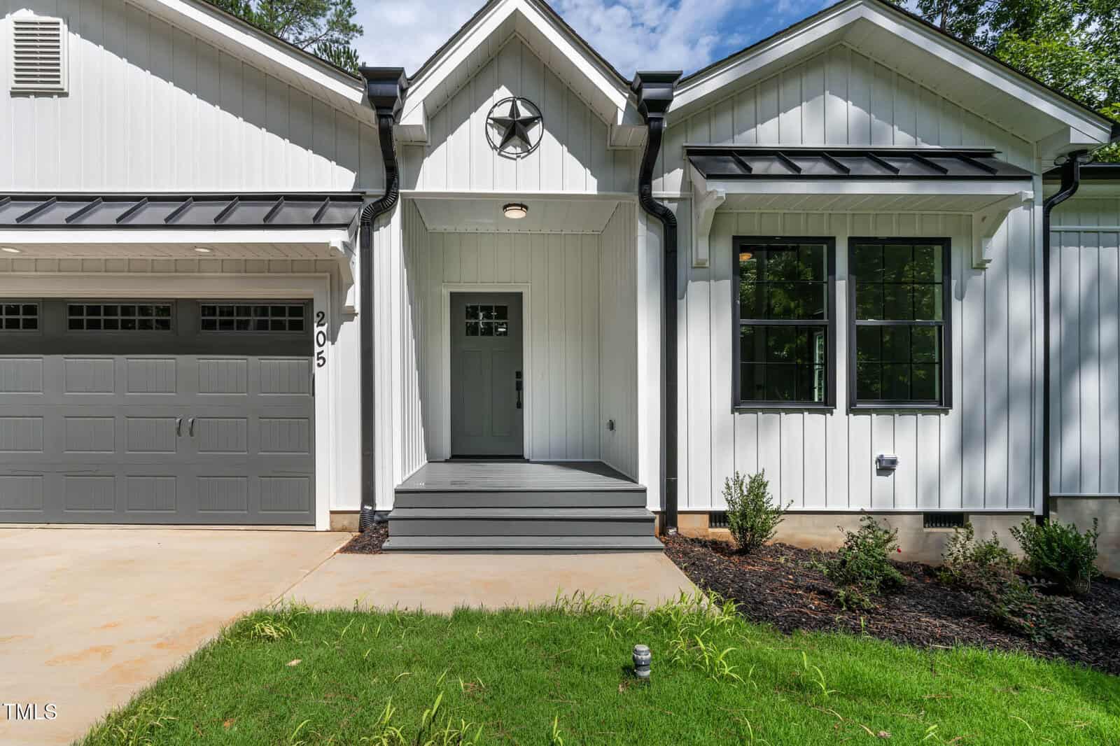 A modern white house with black trim, a gray front door, covered porch with steps, garage on the left, and black-framed windows. Small shrubs and fresh landscaping border the entrance.