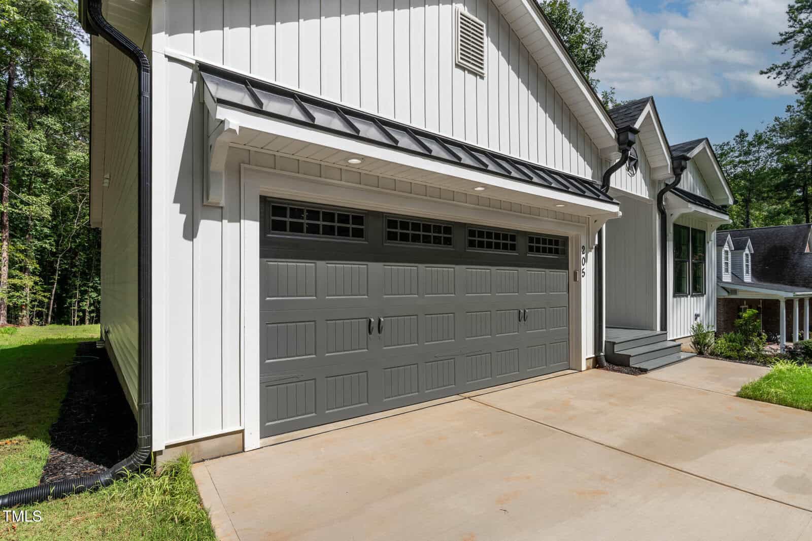 A modern house exterior featuring white vertical siding and a double gray garage door with rectangular windows, a concrete driveway, and some greenery on the side.
