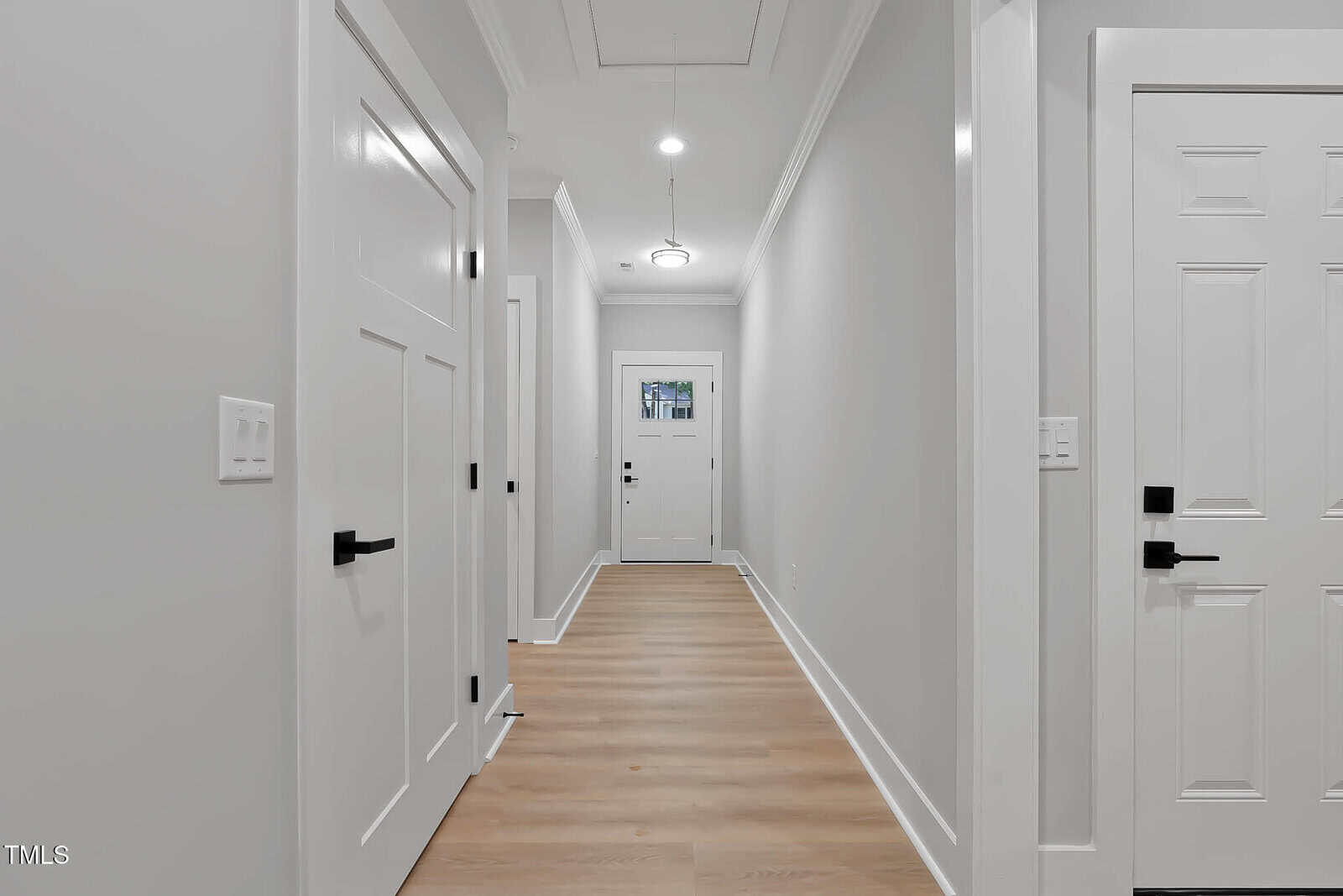 A bright, modern hallway with light wood floors, white walls, and several white doors. A pendant light hangs from the ceiling, and natural light enters through a small window above the door at the end.