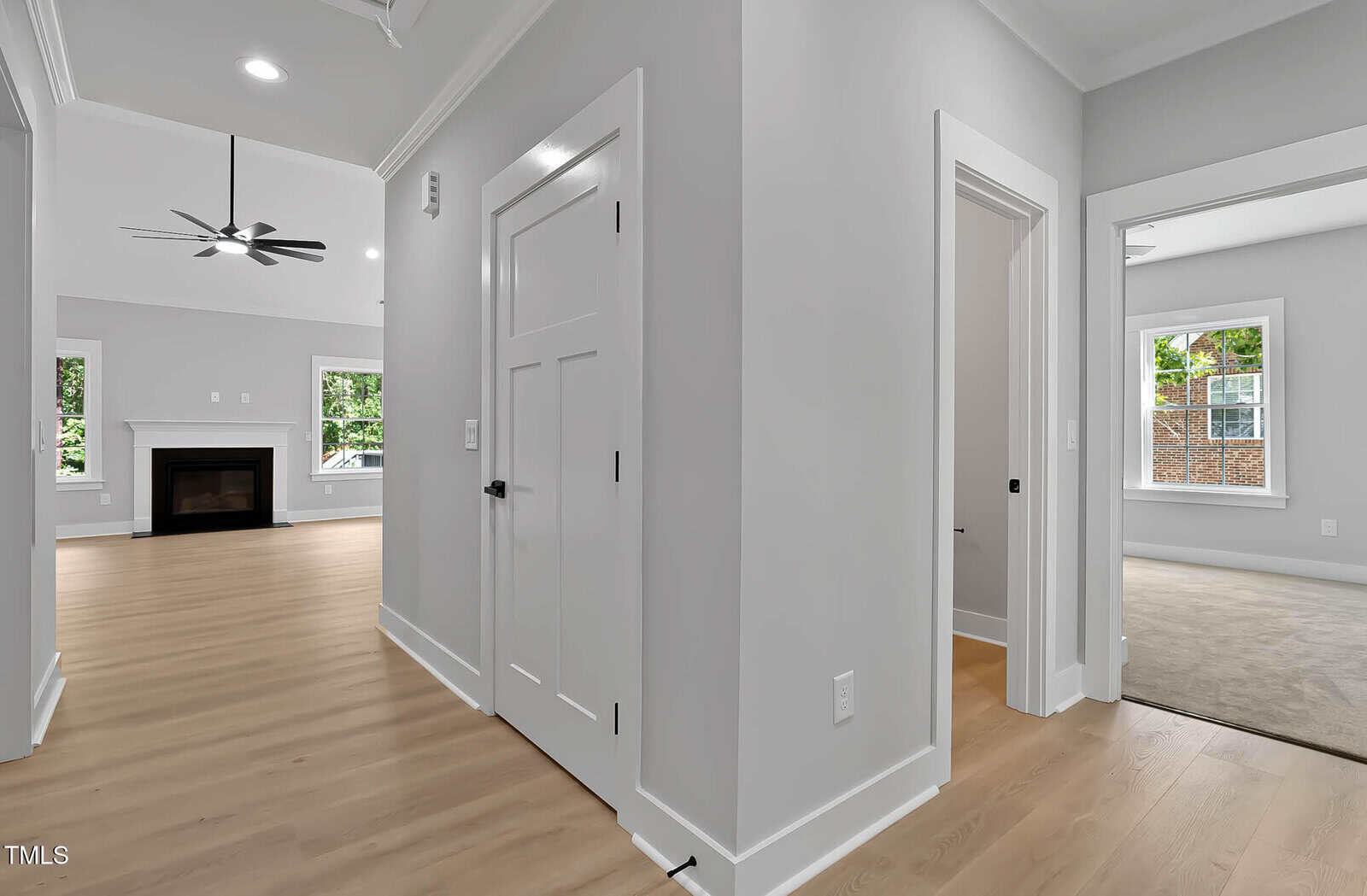 Hallway with light wood flooring, white walls, and white trim. Doors lead to different rooms, including a living area with a fireplace and ceiling fan, and a carpeted bedroom. Large windows provide natural light.