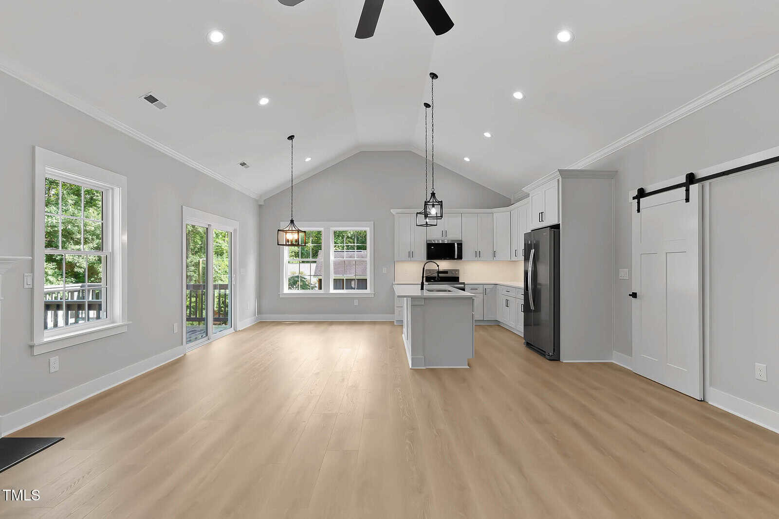 Open-concept kitchen and dining area with light wood floors, white cabinets, a central island, stainless steel appliances, and sliding barn door. Large windows and glass door let in natural light. Ceiling lights and fans are visible.
