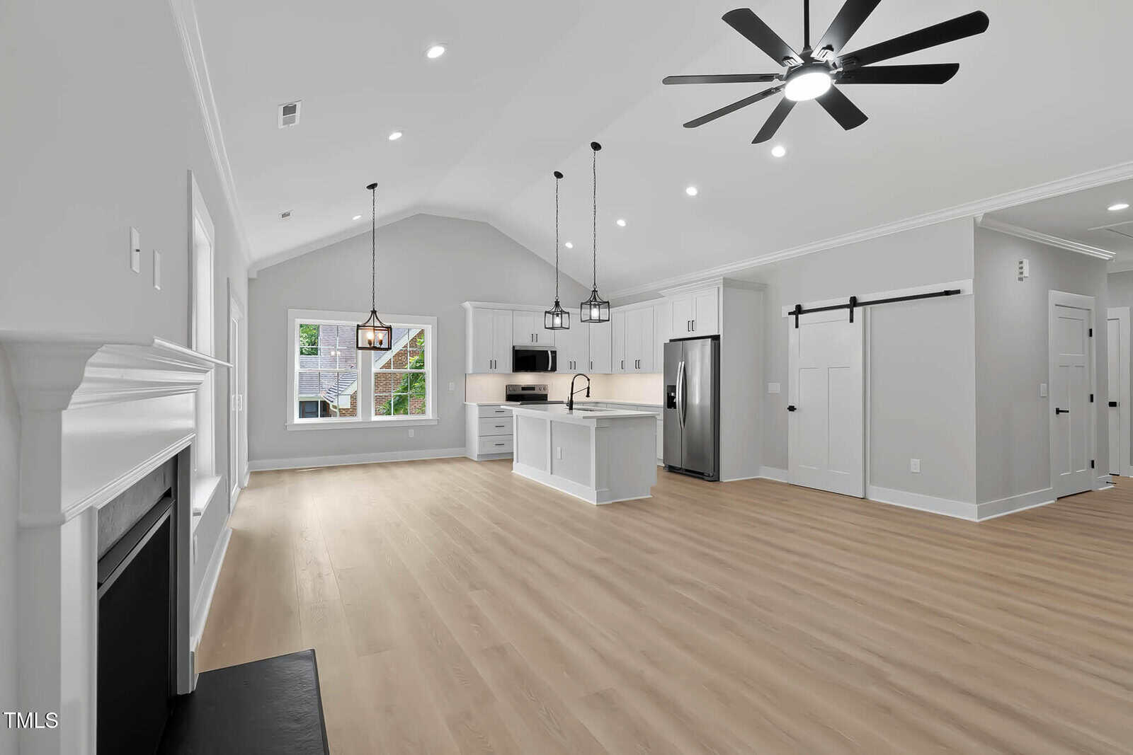 Open-concept kitchen and living area with light wood flooring, white cabinets, stainless steel appliances, a central island, ceiling fan, fireplace, large window, and a sliding barn door leading to another room.