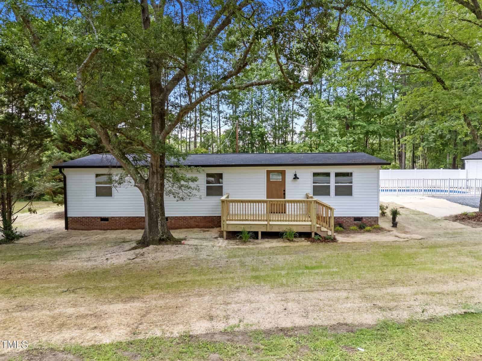 A single-story white house with a brick foundation, small wooden front porch, and a large tree in the front yard sits at 1527 Cash Road in Creedmoor, surrounded by grass, trees, and a wooded area in the background.