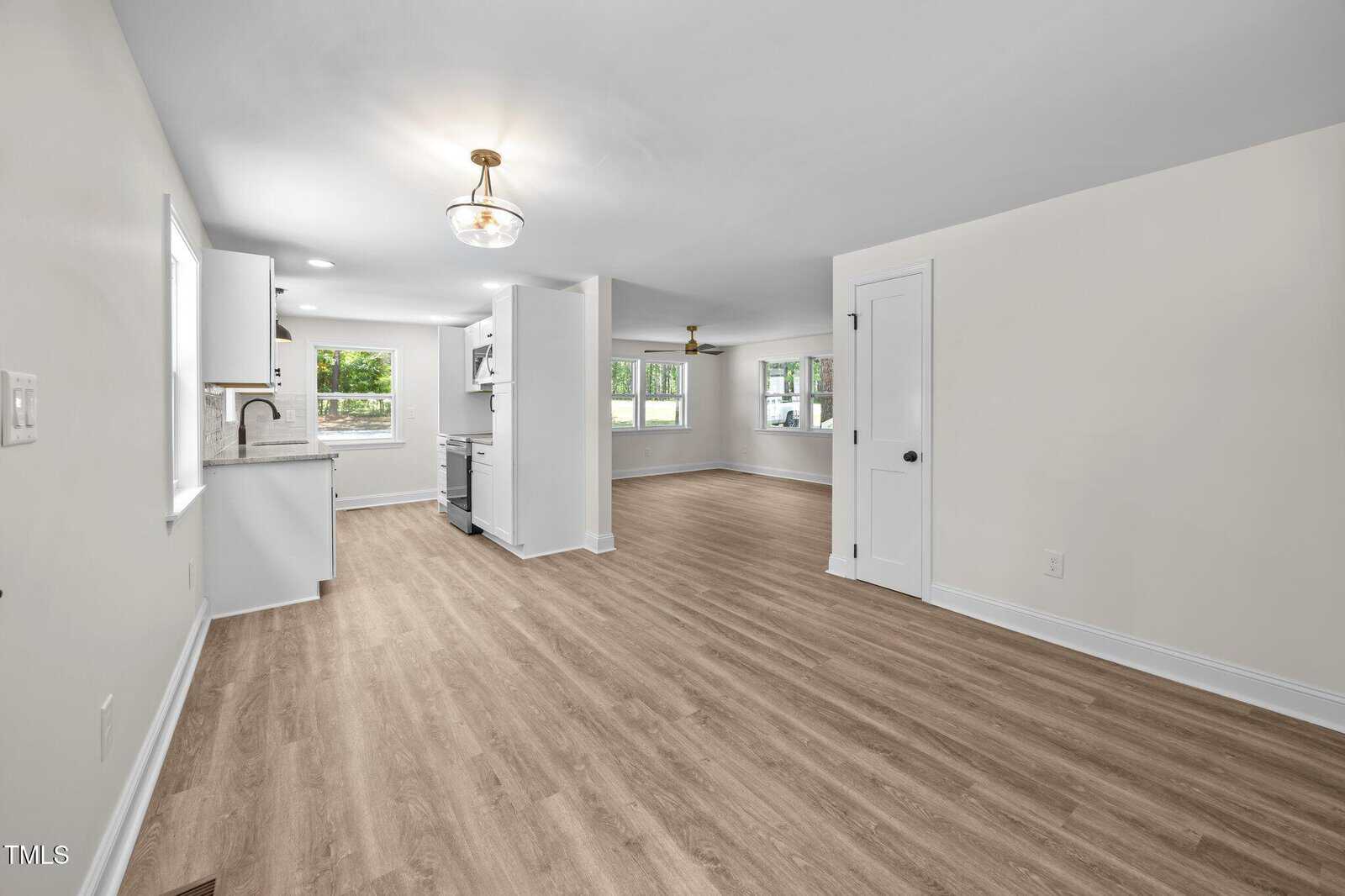 Bright, open-concept living space with light wood flooring, white walls, and modern lighting. The area includes a kitchen with white cabinets and a stainless steel stove, and a living room with multiple windows.