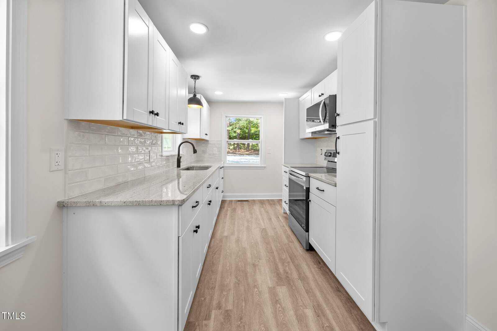 A modern galley kitchen with white cabinets, stainless steel appliances, light stone countertops, a subway tile backsplash, wood-style flooring, and a window at the end of the room letting in natural light.