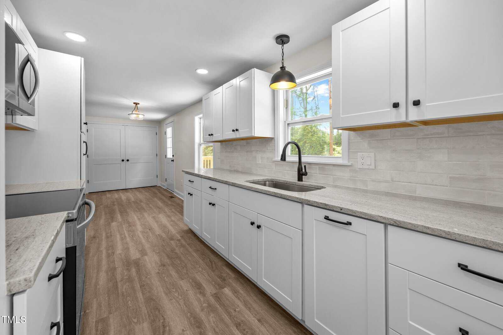 A modern galley kitchen with white cabinets, light gray countertops, stainless steel appliances, a black faucet, tile backsplash, wood-look flooring, and large windows letting in natural light.