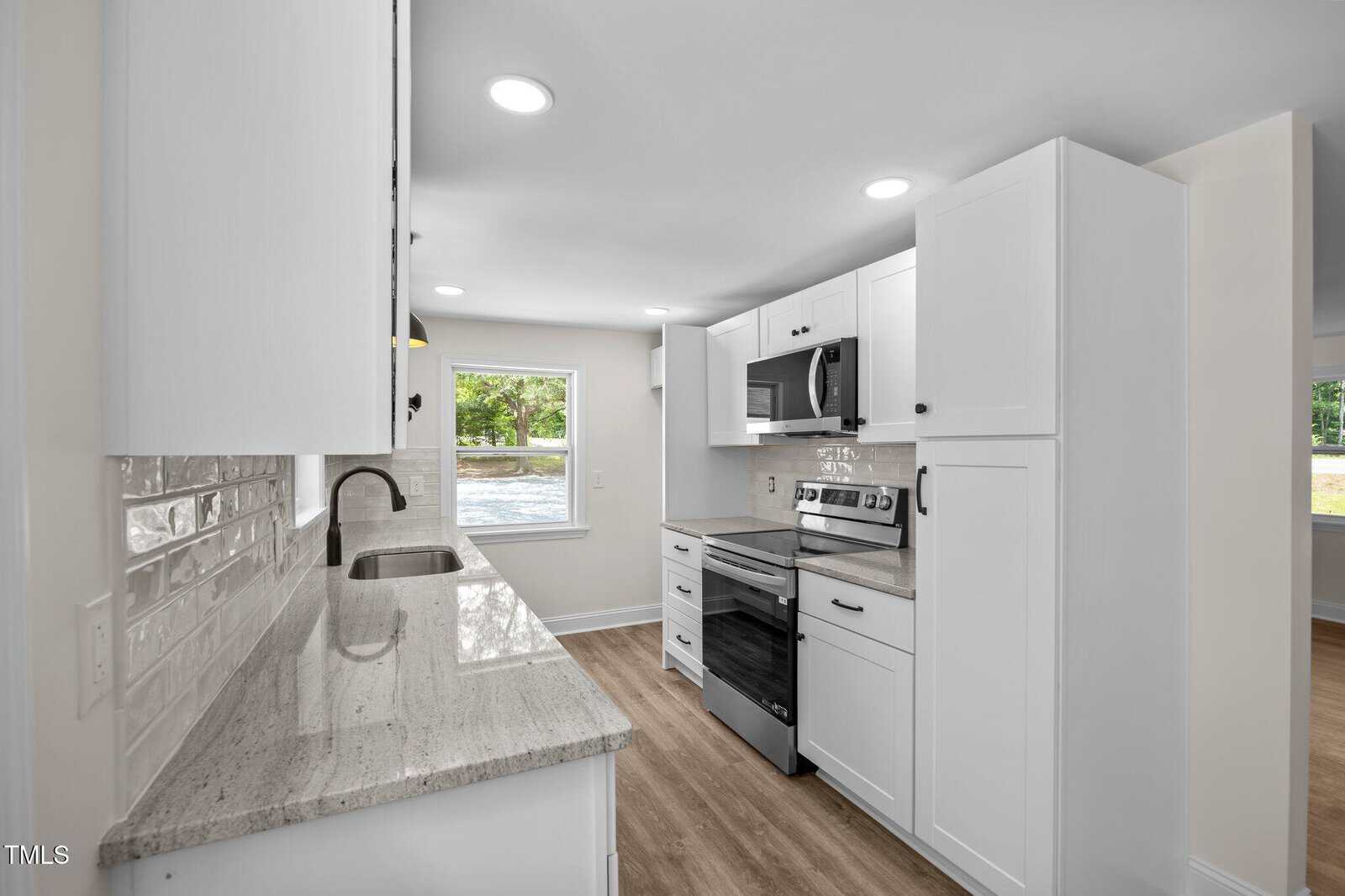 A modern kitchen with white cabinets, stainless steel appliances, light granite countertops, a white subway tile backsplash, and wood-look flooring. Large windows provide natural light and views of greenery outside.