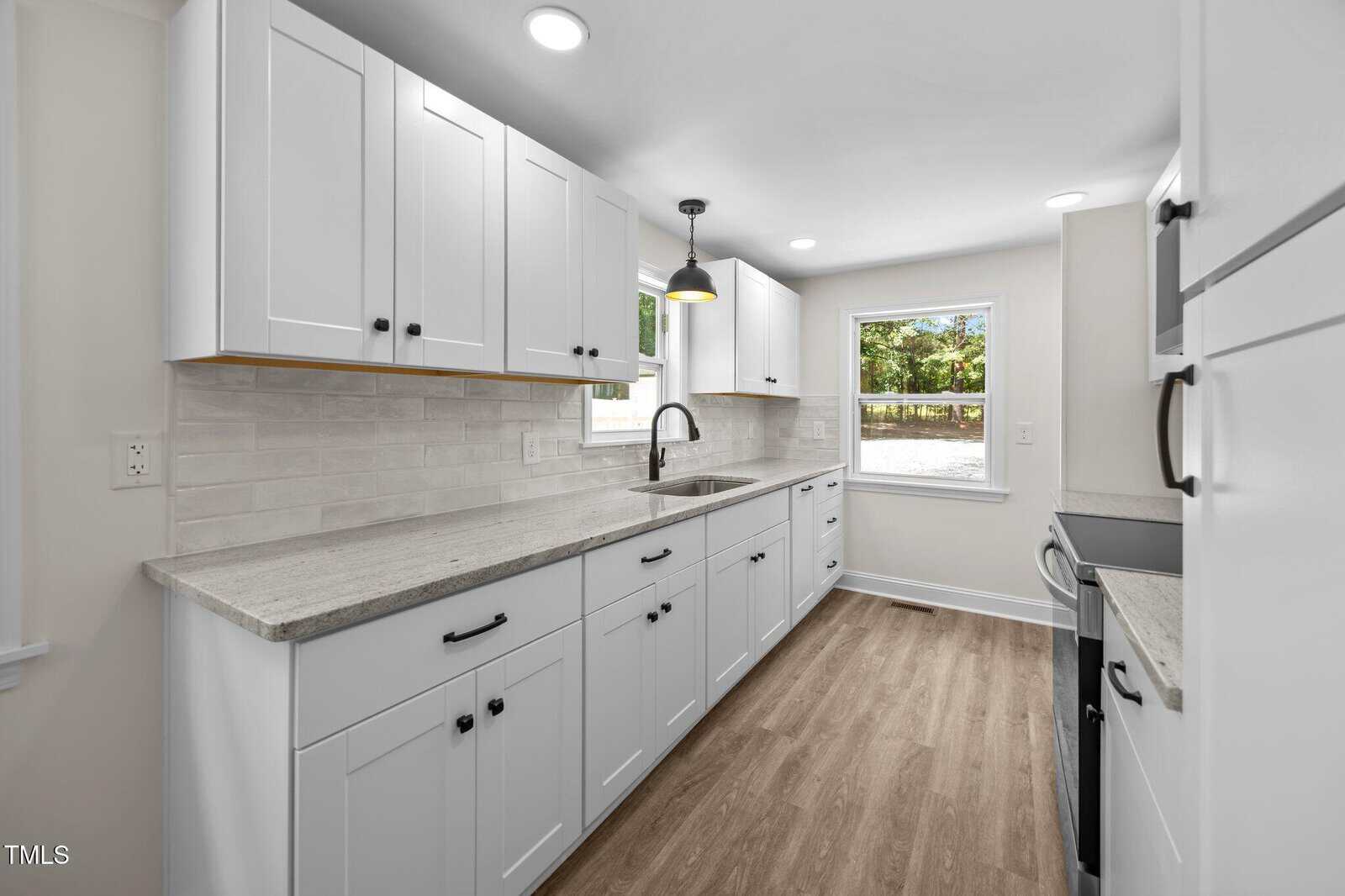A modern kitchen with white cabinets, light gray countertops, a stainless steel sink under a window, light wood flooring, white subway tile backsplash, and built-in lighting on the ceiling.