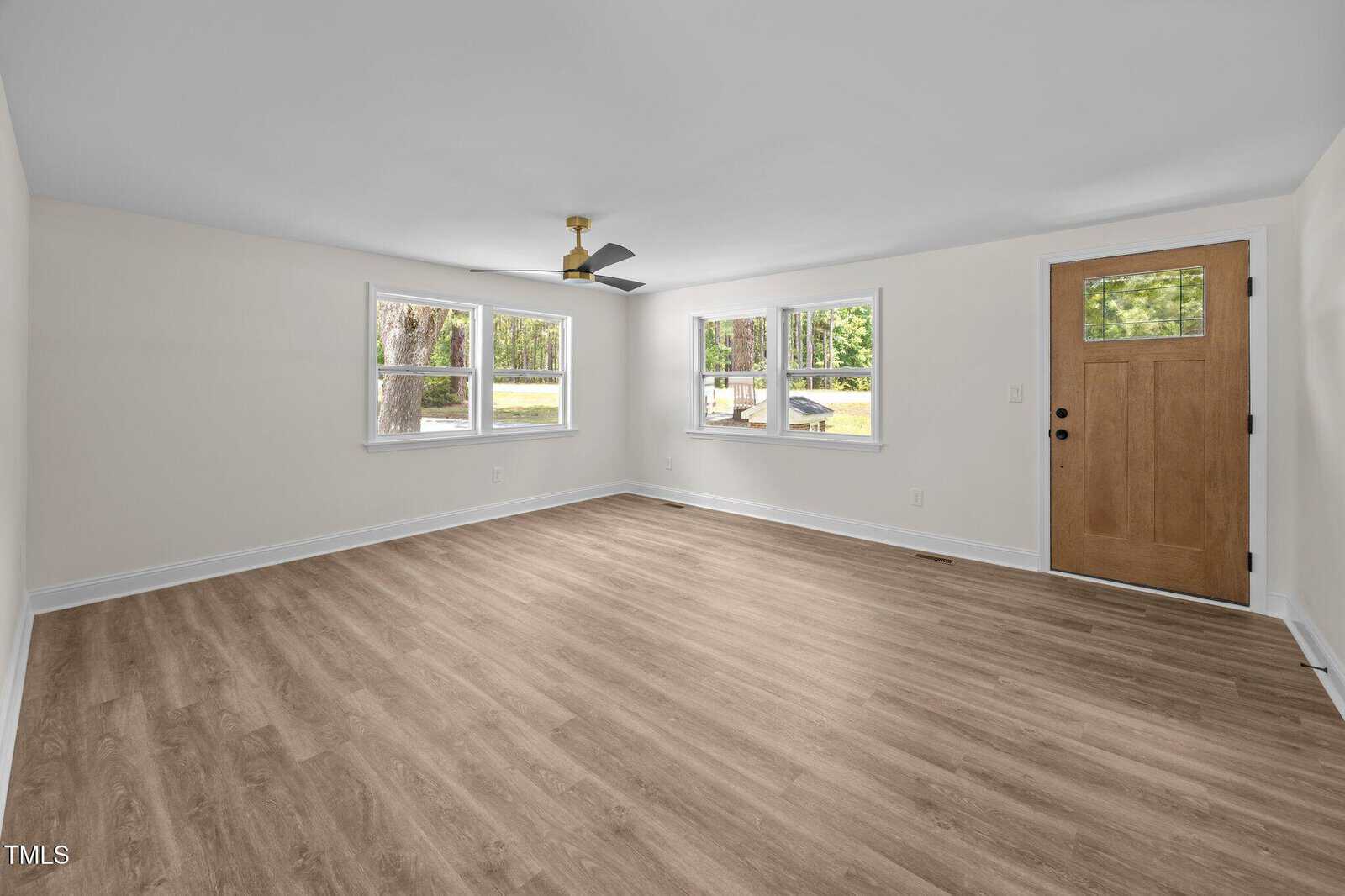 Empty living room with light-colored walls, wood-style flooring, a ceiling fan with black blades, three windows, and a wooden door with a small square window. Natural light is coming through the windows.