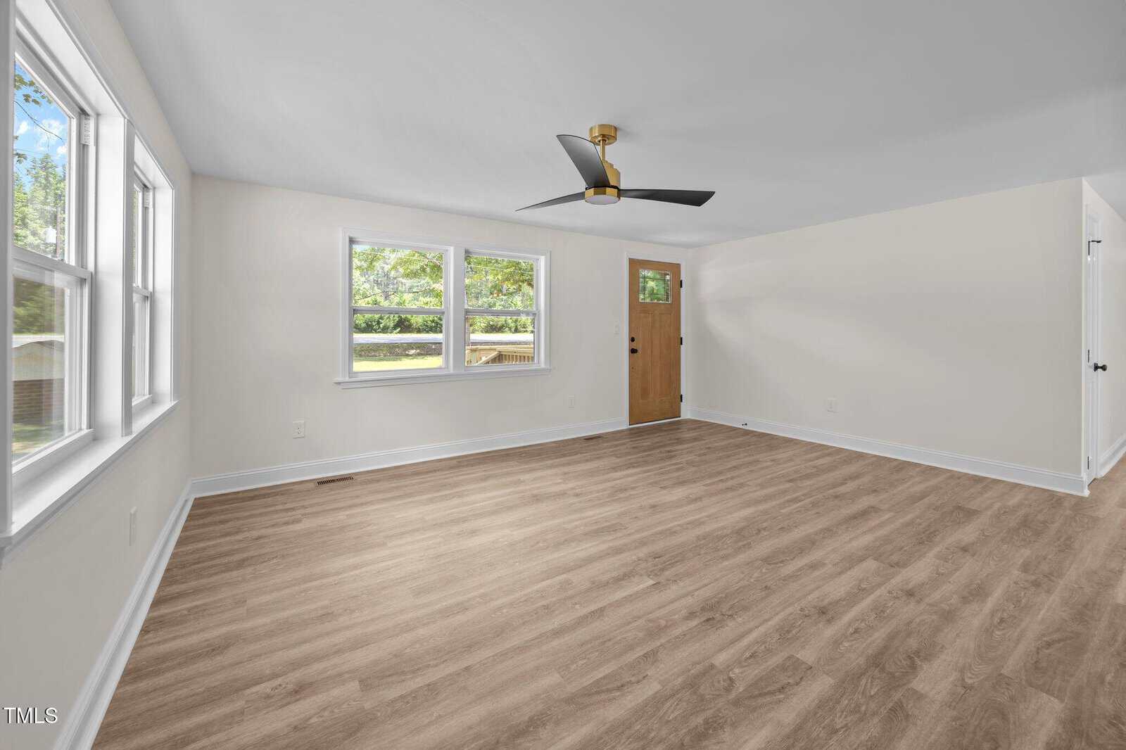 A bright, empty room with light wood flooring, white walls, a ceiling fan with black blades, three large windows, and a wooden door leading outside. Daylight streams in through the windows.