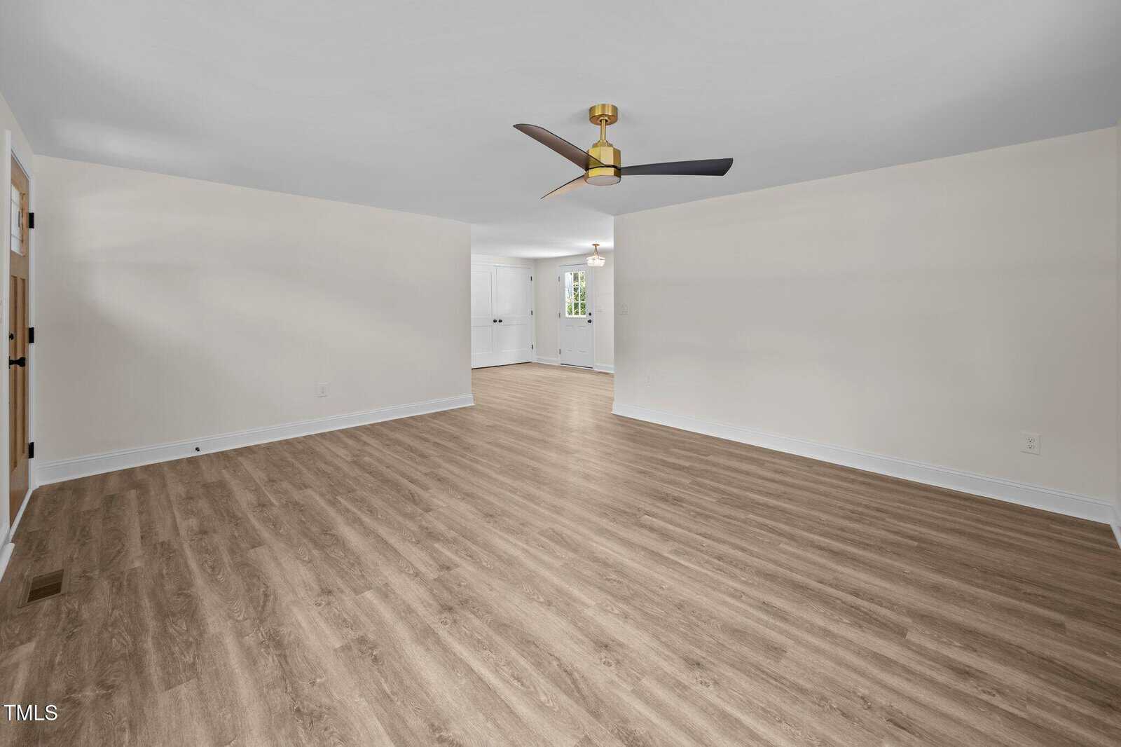 A spacious, empty room with light wood flooring, cream-colored walls, a modern black and gold ceiling fan, and an open layout leading to another area with white cabinetry and a door.