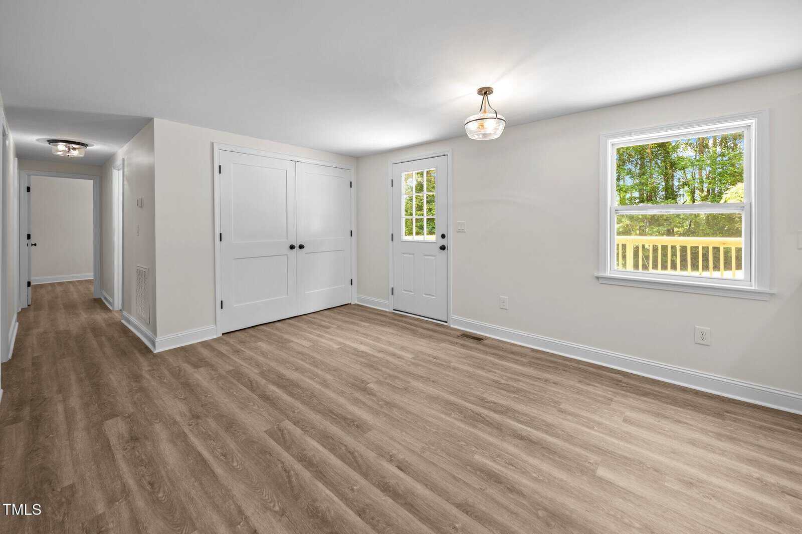 A bright, empty room with wood-look flooring, white walls, a ceiling light fixture, two closed white double doors, a single door with a window, and a window showing green trees outside.