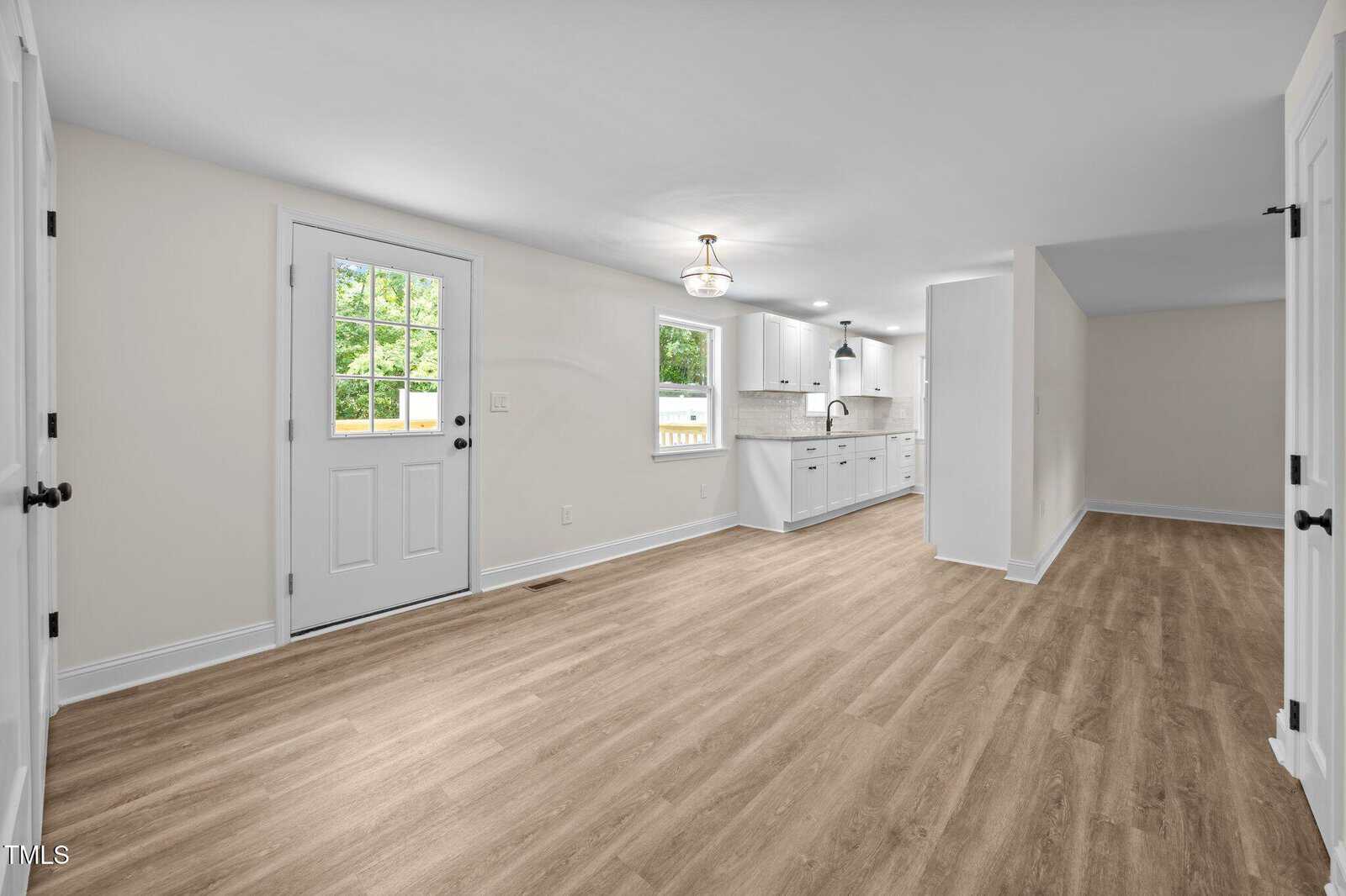 Bright open room with light wood floors, white walls, a door and window on the left, kitchen with white cabinets and sink in the back, modern light fixture on the ceiling.