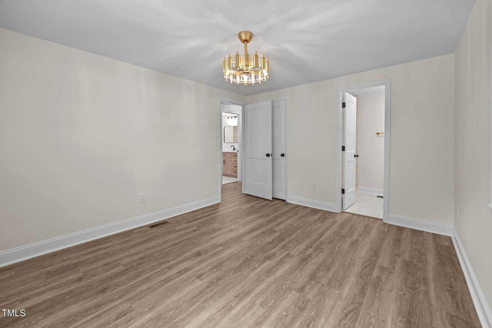 A bright, empty room with light beige walls, wood-look flooring, a gold chandelier, and two white doors leading to a bathroom and a closet.