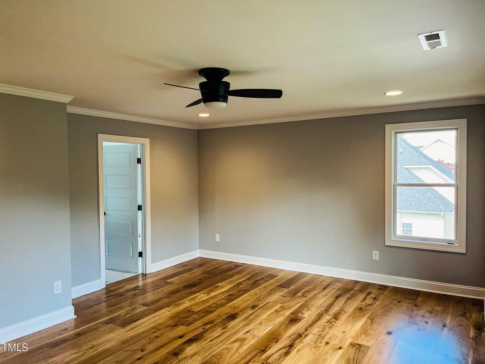 A bright, empty room with light gray walls, wood floors, a black ceiling fan, two windows, and a white door leading to another room. White baseboards and crown molding are visible along the edges.