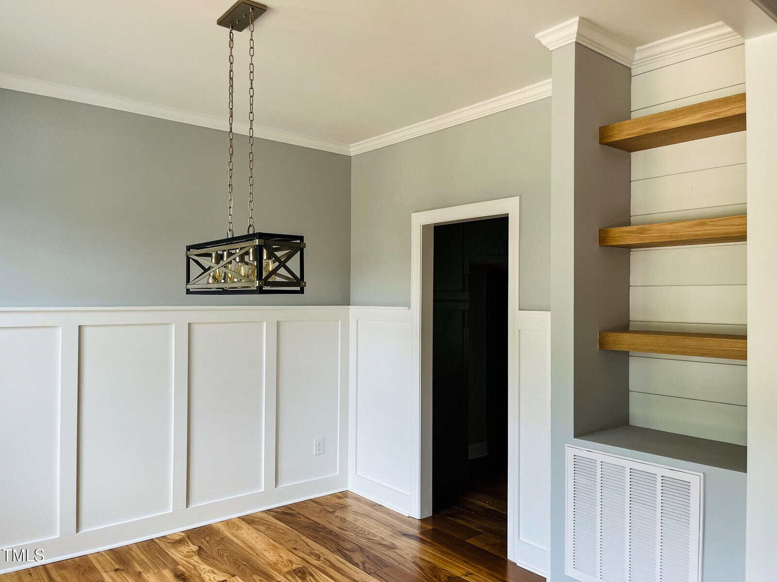 A room with light gray walls, white wainscoting, wood floors, a black metal chandelier, a doorway leading to a dark room, and open wooden shelves built into the corner above a vent.