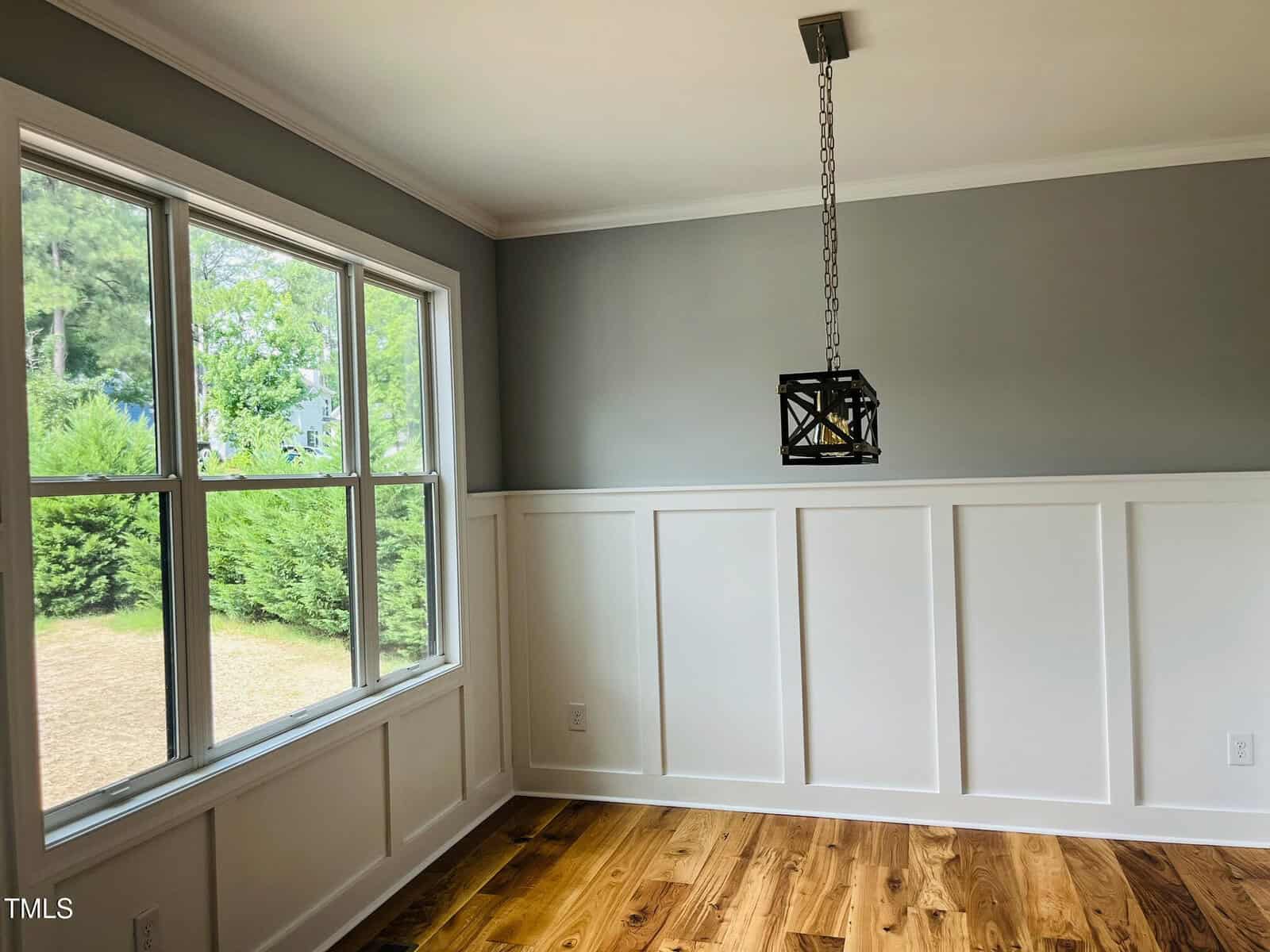 A bright, empty room with gray upper walls, white wainscoting, hardwood floors, a large window with a view of greenery outside, and a modern black pendant light hanging from the ceiling.