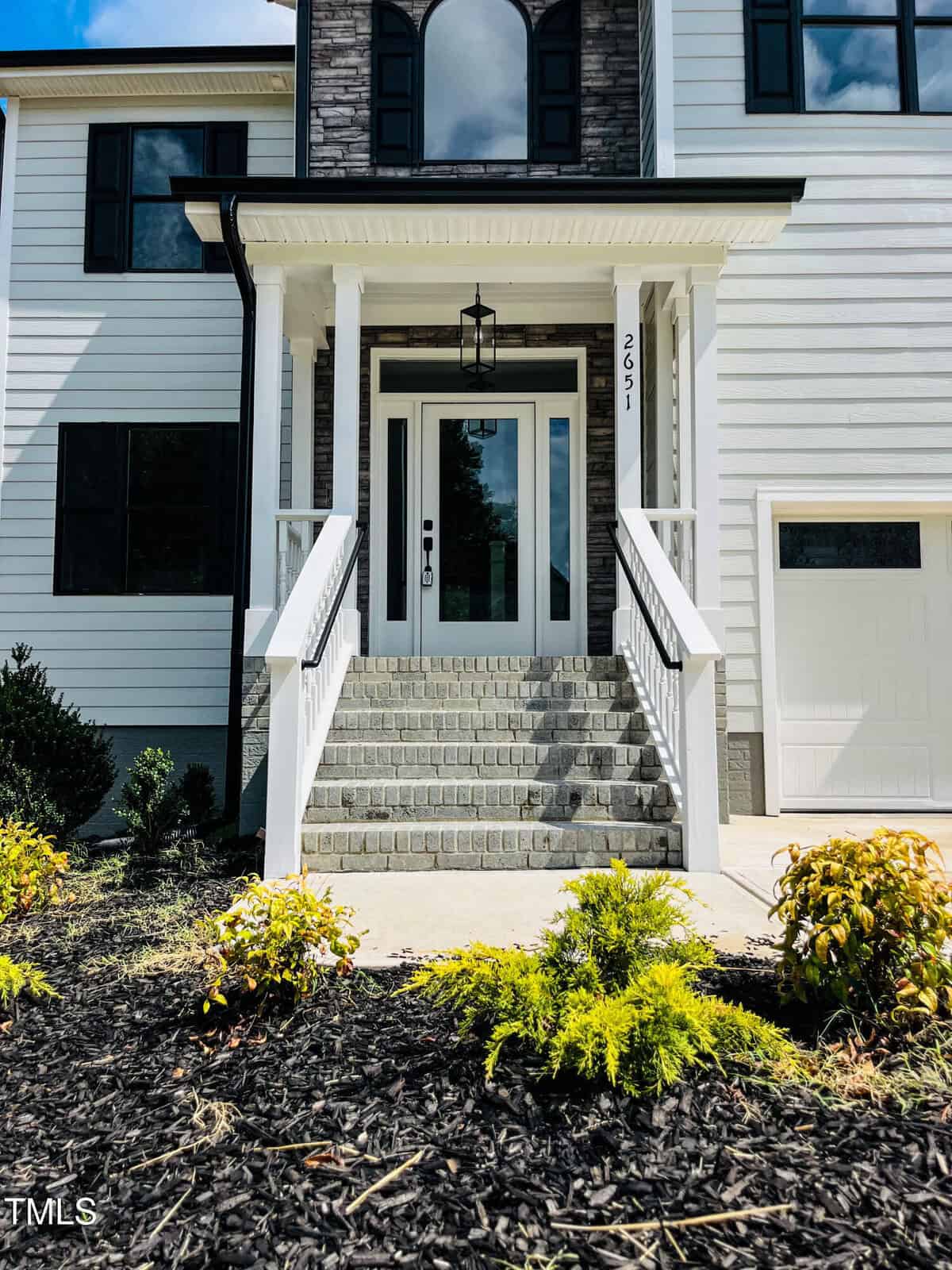 Front view of a house with a white exterior, stone steps leading up to a glass front door, two railings, black-trimmed windows, a small garden with shrubs, and part of a garage door visible on the right.