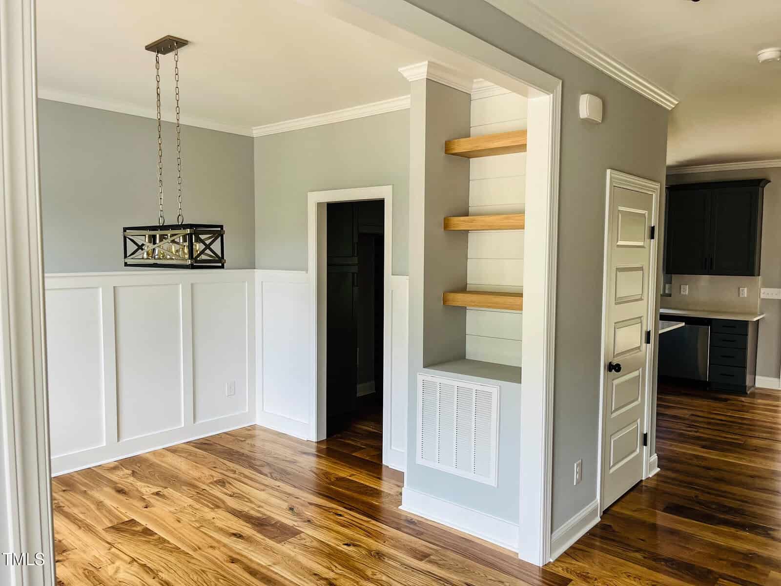 A dining area with hardwood floors, white wainscoting, and gray walls. Built-in wooden shelves are on the right, and a modern rectangular light fixture hangs above. A hallway and kitchen are visible in the background.