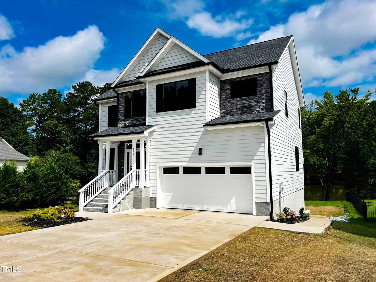 A two-story white house with black trim features a covered front porch, double garage, and concrete driveway. The yard is landscaped with shrubs, and trees surround the property under a partly cloudy sky.