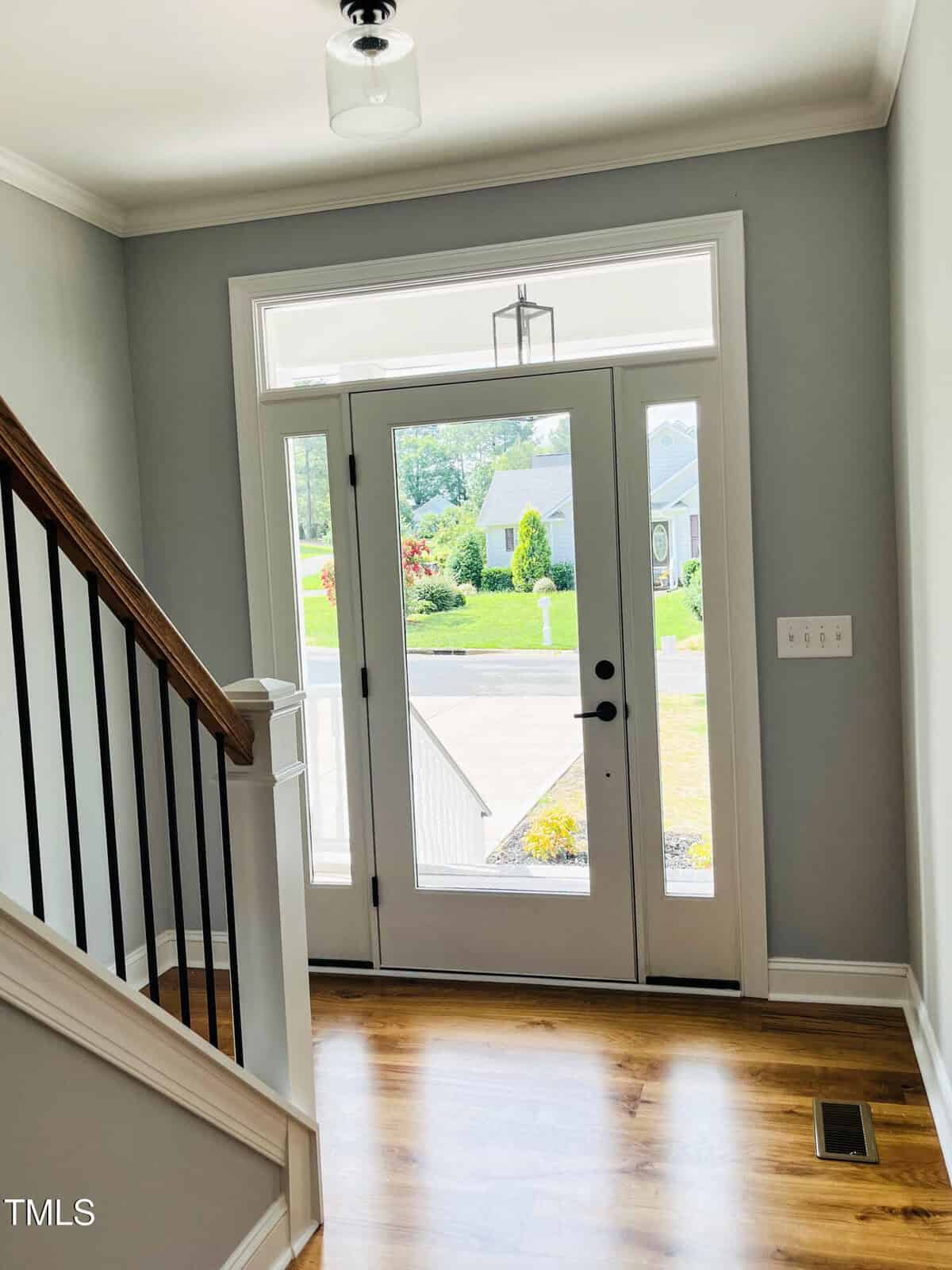 A bright entryway with a glass front door and side windows, hardwood floors, gray walls, white trim, a staircase with dark handrails, and a view of the street and greenery outside.