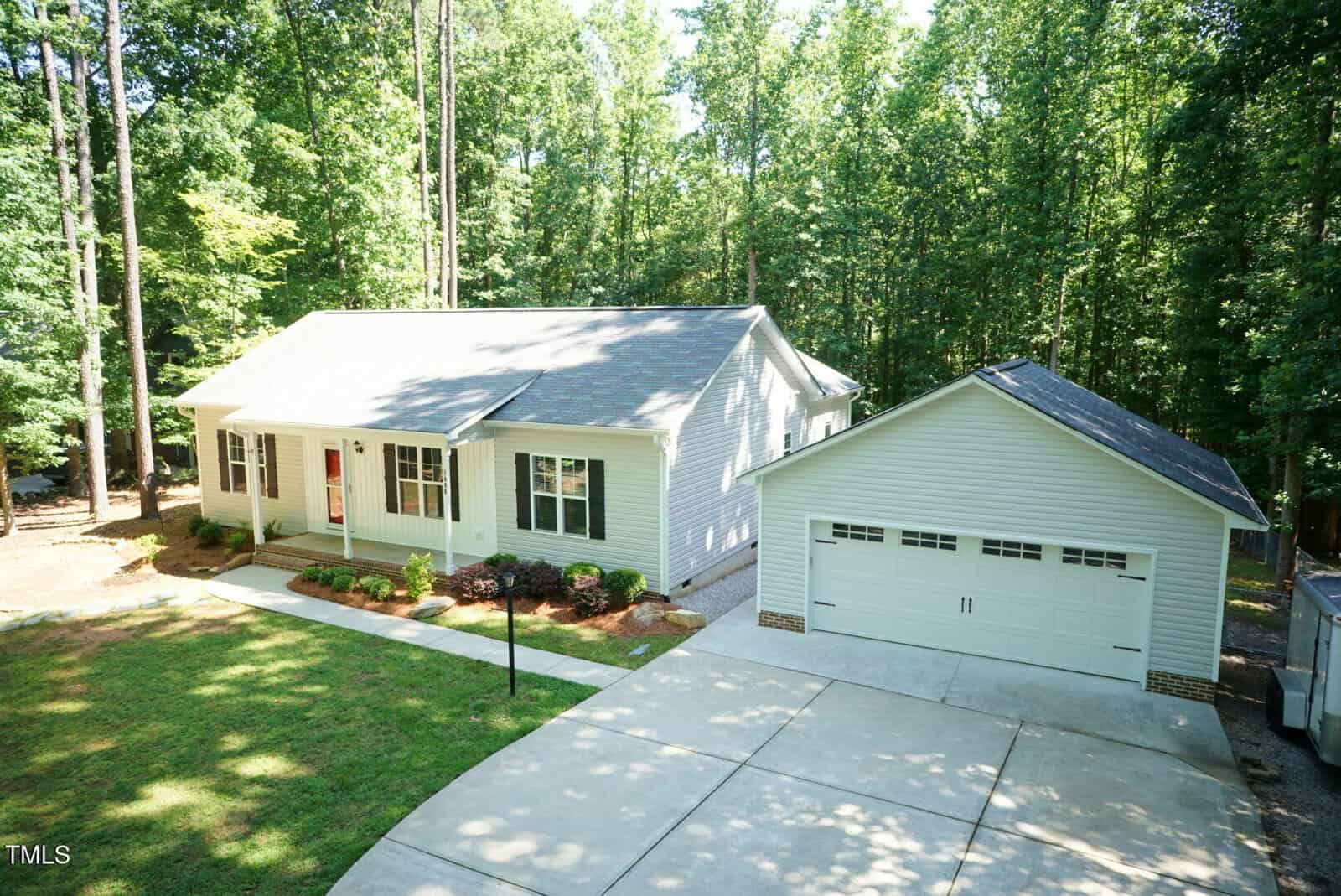 Single-story white house with a front porch and detached two-car garage, surrounded by trees. A wide driveway leads to the garage, and the lawn and landscaping are neatly maintained.