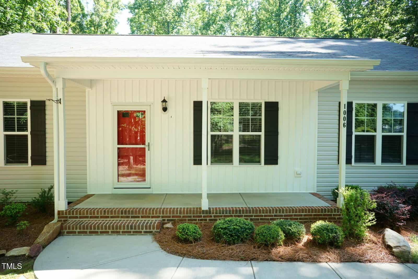 A single-story house with white siding, a small covered front porch, a red front door with a glass storm door, brick steps, and neatly trimmed bushes along the walkway.