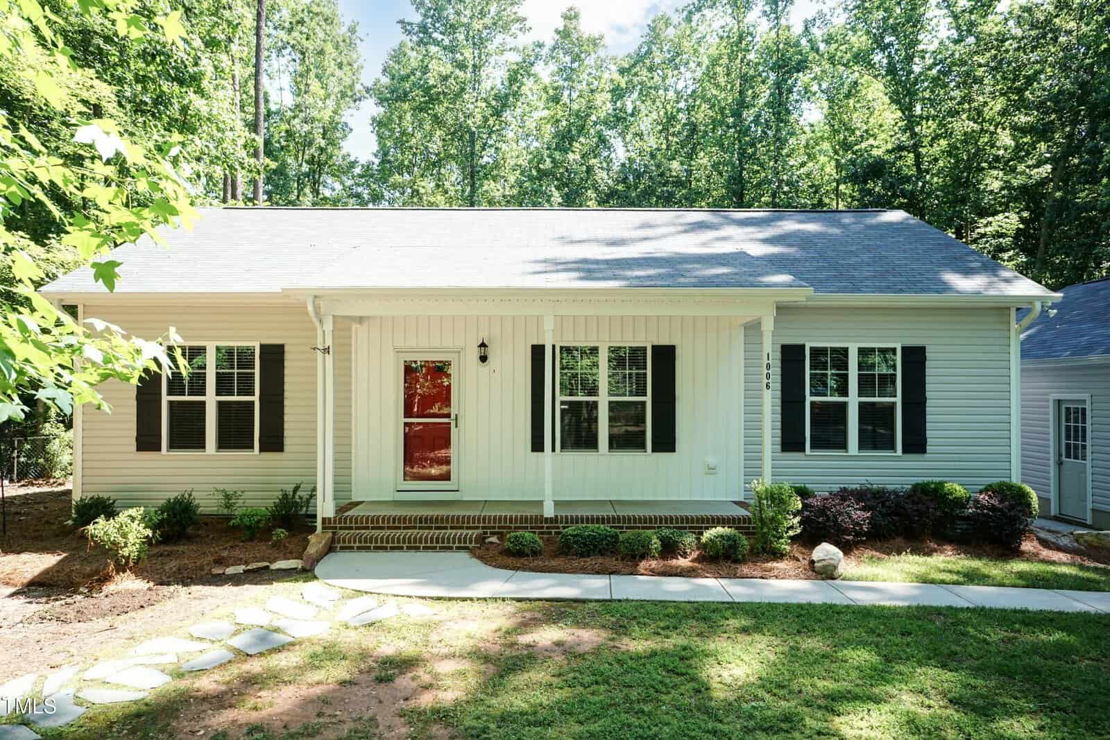 Charming single-story white house at 1006 Oxford Ridge Road in Wake Forest, featuring black shutters, a red front door, a cozy front porch with brick steps, and a landscaped yard bordered by trees and shrubs under bright daylight.