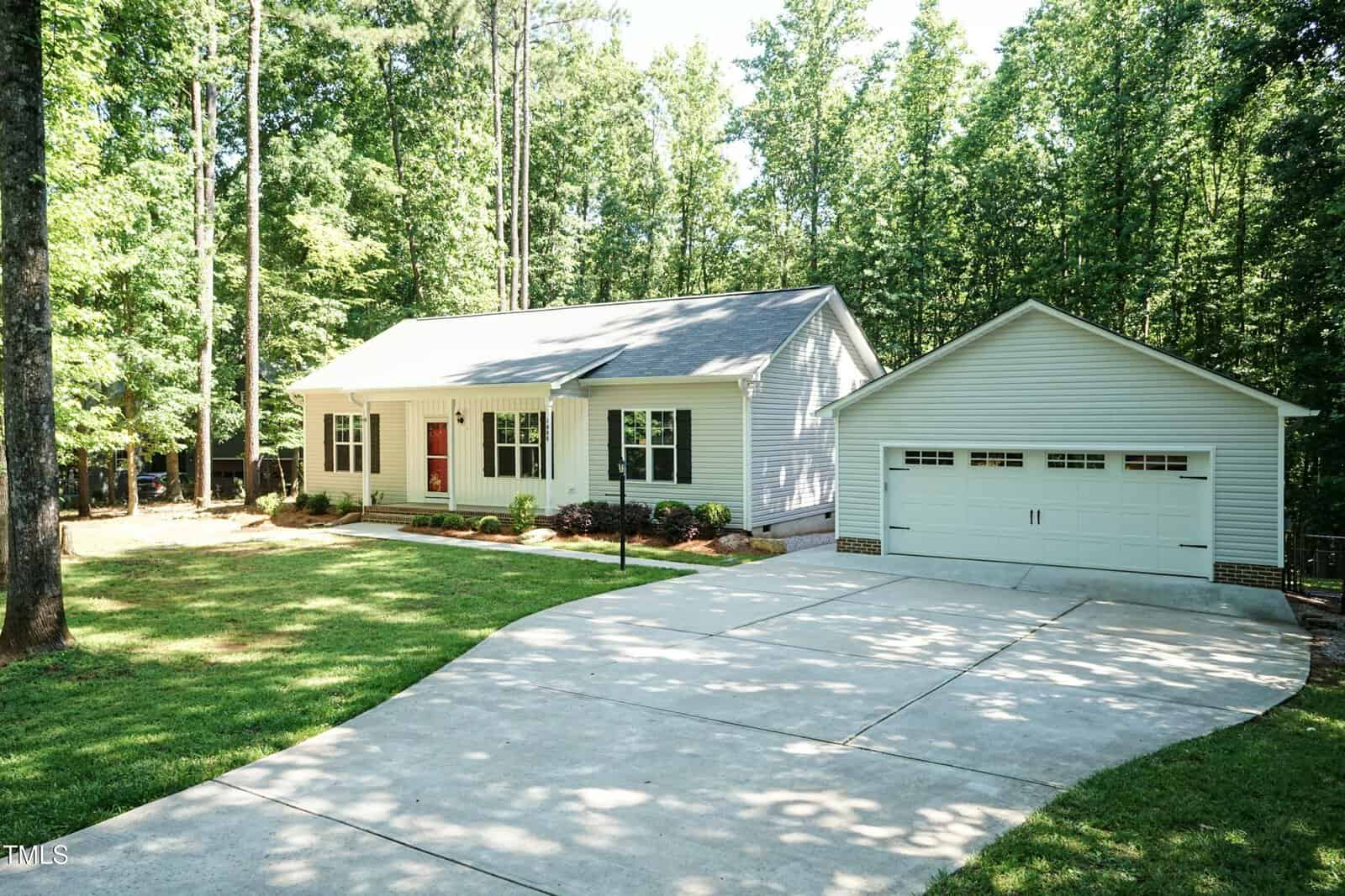 A single-story light gray house with a front porch and red door sits next to a detached two-car garage. The home is surrounded by tall trees and has a wide concrete driveway and a well-maintained lawn.
