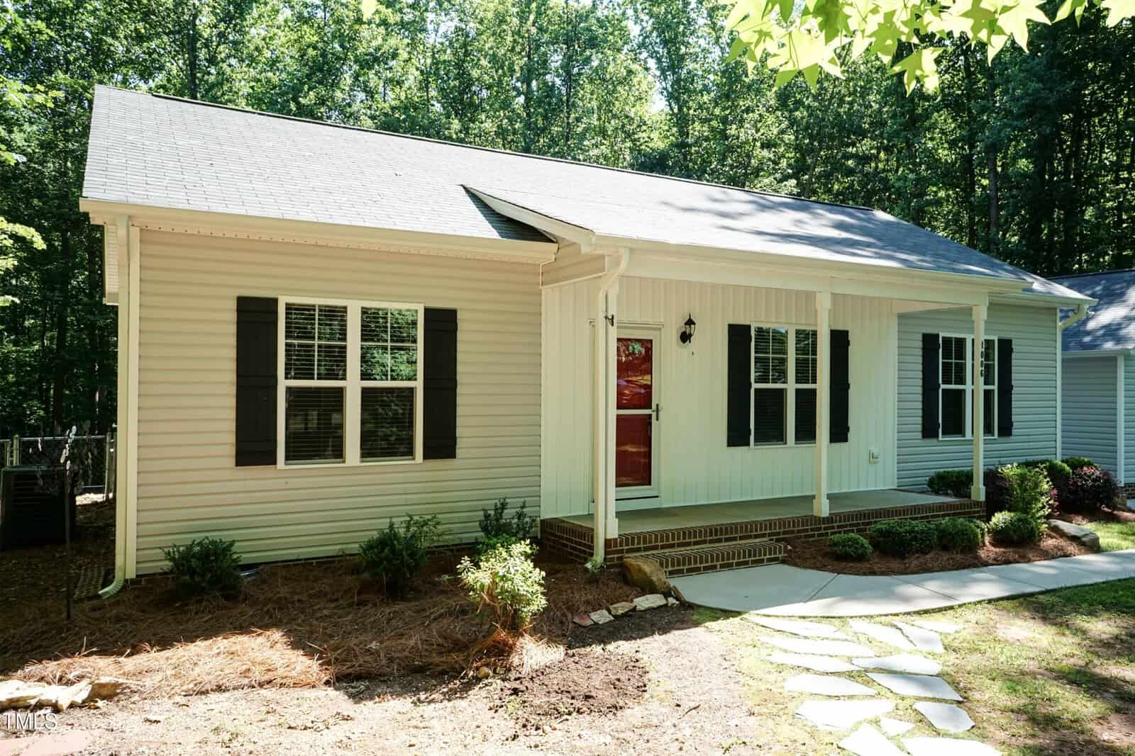 Single-story house with light-colored siding, a dark roof, black shutters, and a red front door. There is a small front porch with steps, simple landscaping, and a stone pathway leading to the entrance.