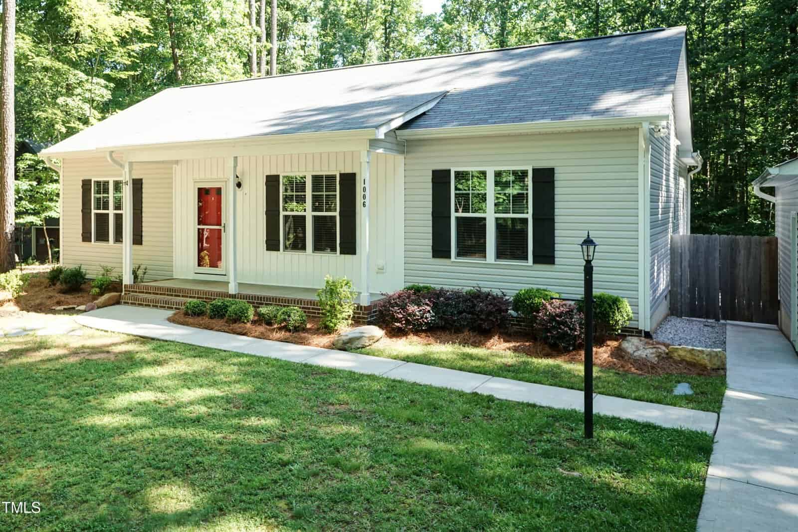 Single-story house with light-colored siding, a gray shingled roof, black shutters, a red front door, a small front porch, landscaped bushes, and a sidewalk leading to the entrance. Surrounded by trees and grass.